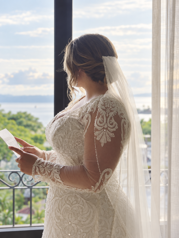 Bride in a white lace wedding dress with a veil, standing on a balcony, reading a card with a view of water and greenery outside.