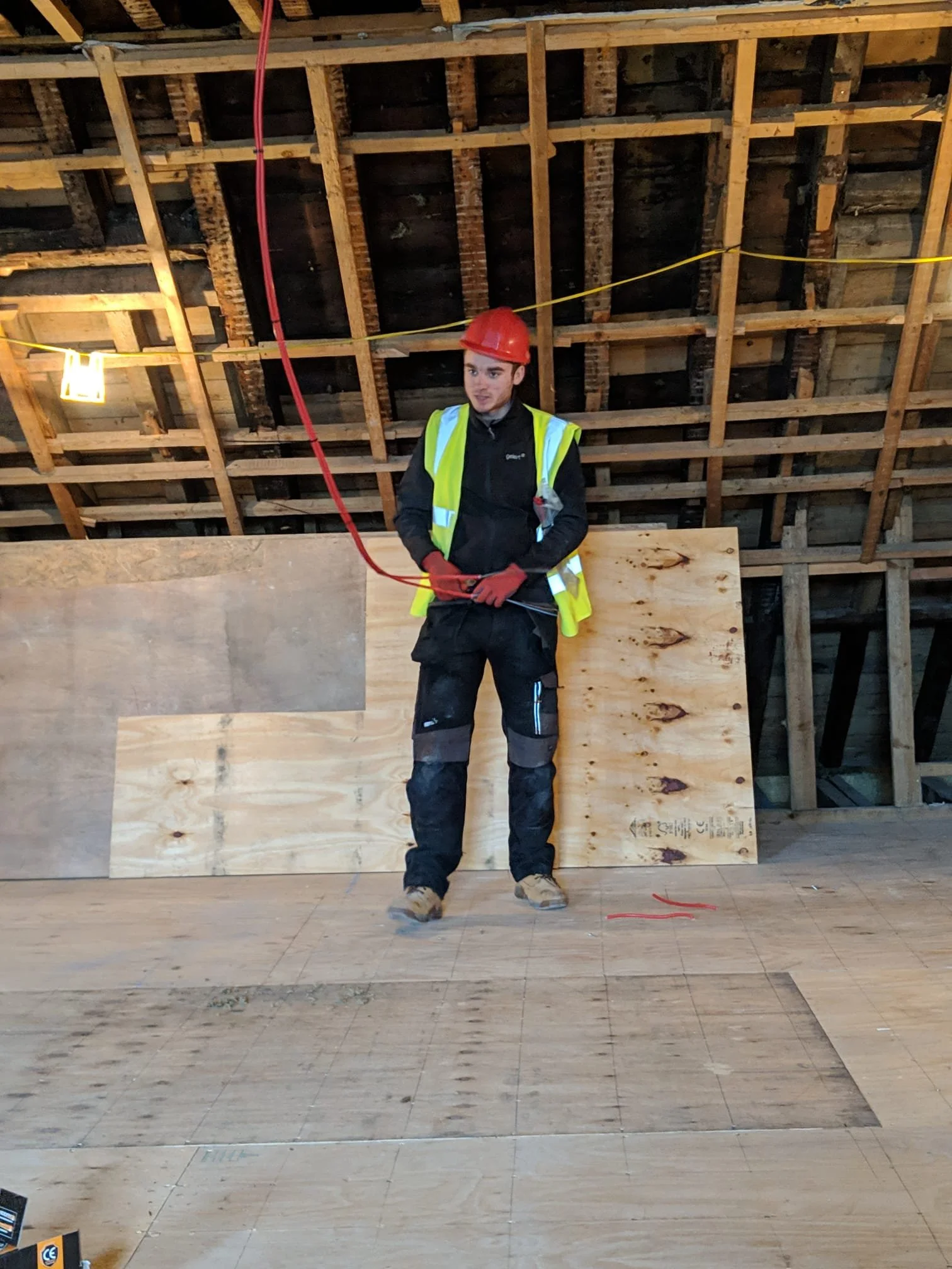 Construction worker wearing a red hard hat, safety vest, and work gloves standing indoors on a plywood platform, holding a red cable. The background shows an unfinished wooden ceiling framework and a yellow electrical wire.
