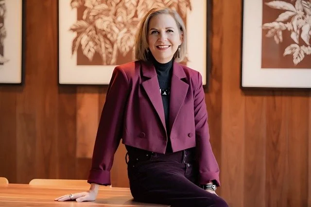 A woman with shoulder-length blonde hair, wearing a maroon blazer and black top, sitting on a wooden table in front of wood-paneled walls with framed botanical artwork.