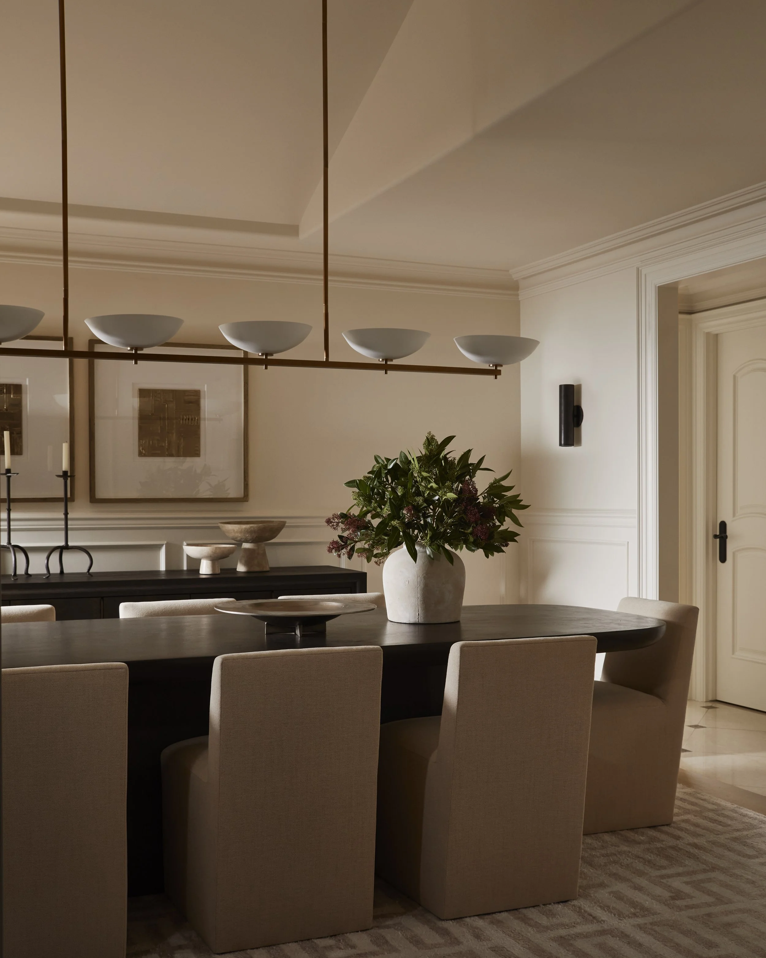 A dining room with a round table surrounded by beige upholstered chairs, a large white vase with green and purple plants on the table, and decorative elements on the wall including framed artwork and a black wall sconce.