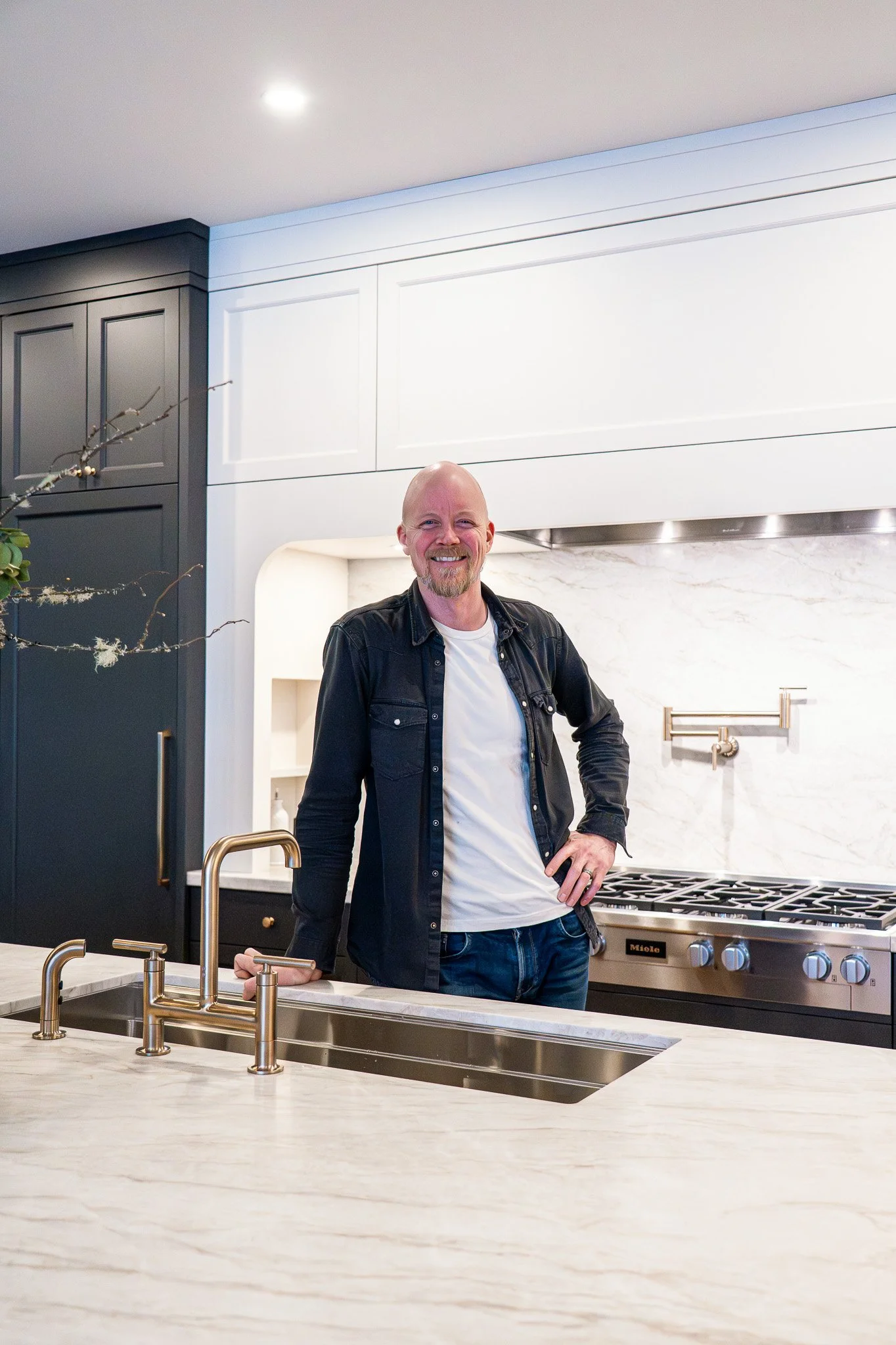 A smiling man with a beard and a shaved head, wearing a black jacket and white T-shirt, standing in a modern kitchen with white marble countertops, black cabinets, and a stainless steel gas stove.