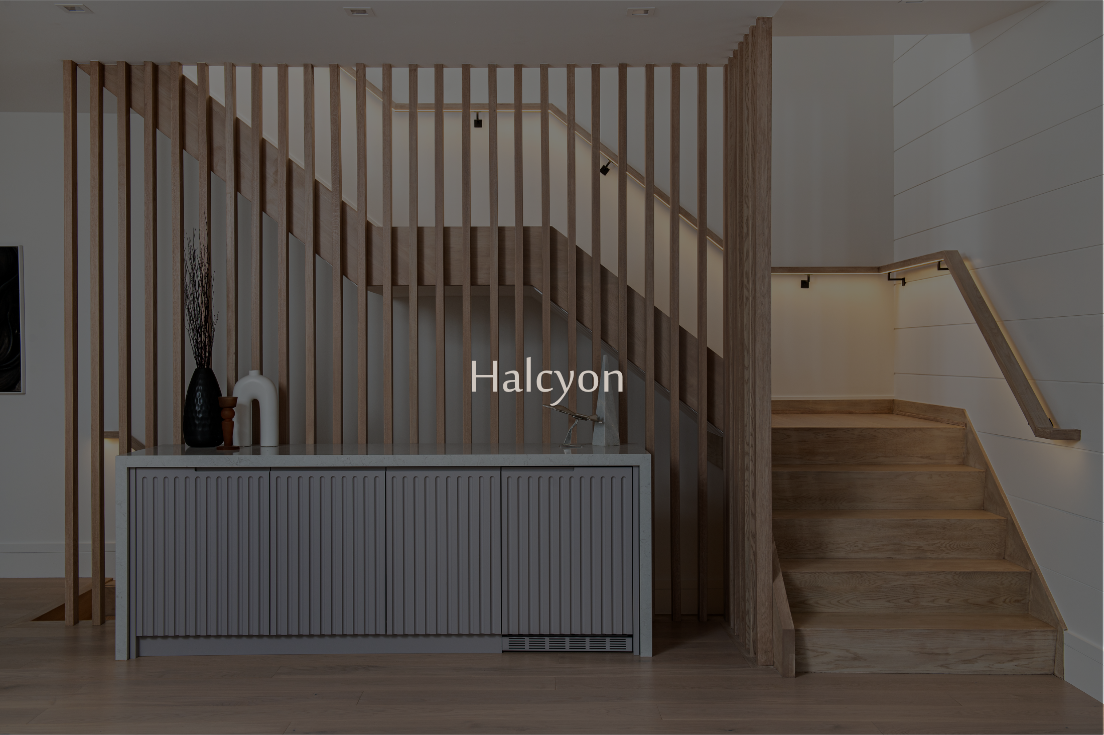 Interior view of a staircase with a wooden handrail and vertical wooden slats, a white cabinet with decorative vases on top, and a wall with horizontal wood paneling, illuminated by subtle lighting.