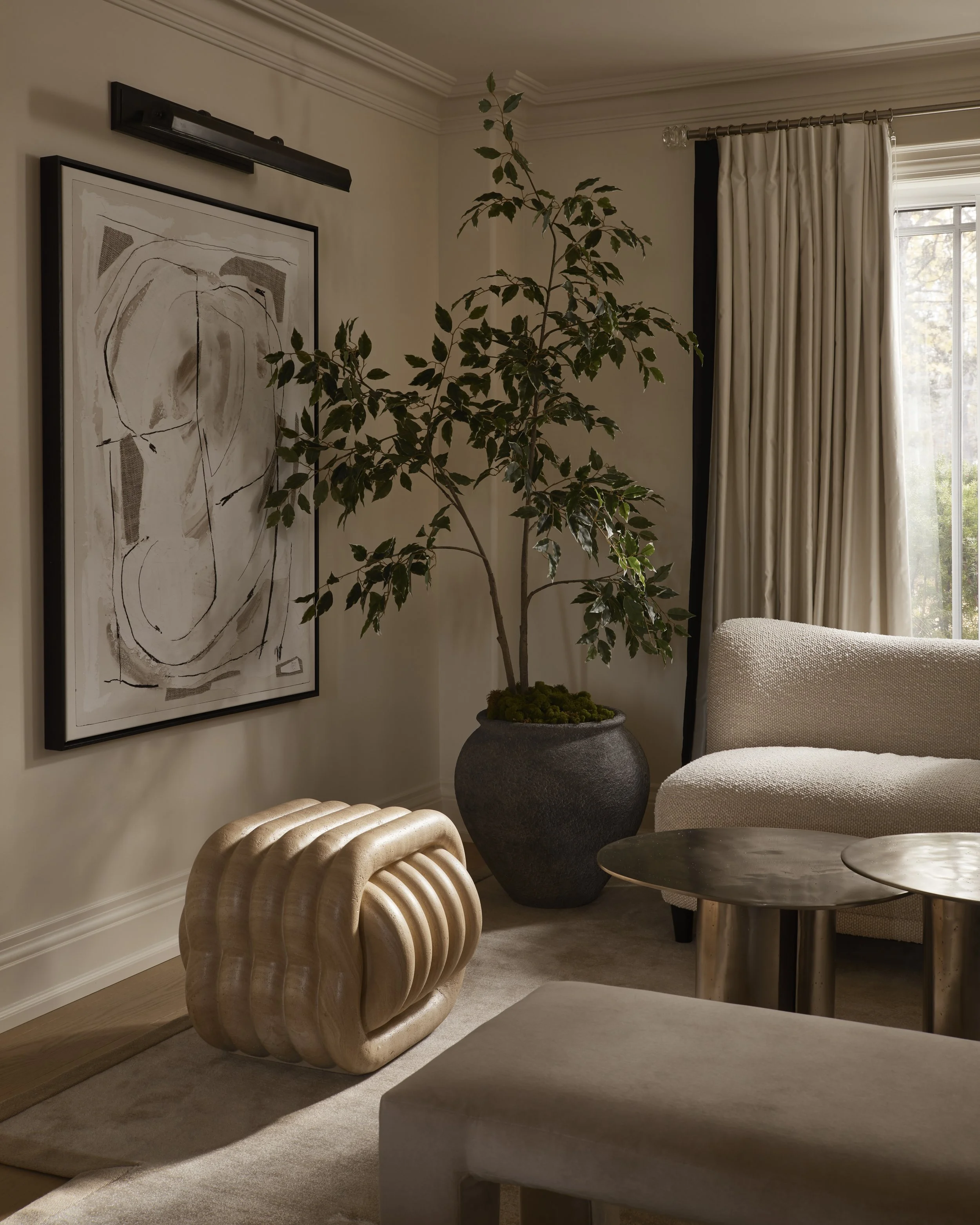 Living room corner with abstract art on wall, large potted plant, and beige furniture, including a cream armchair and wooden coffee table, next to window with cream curtains.