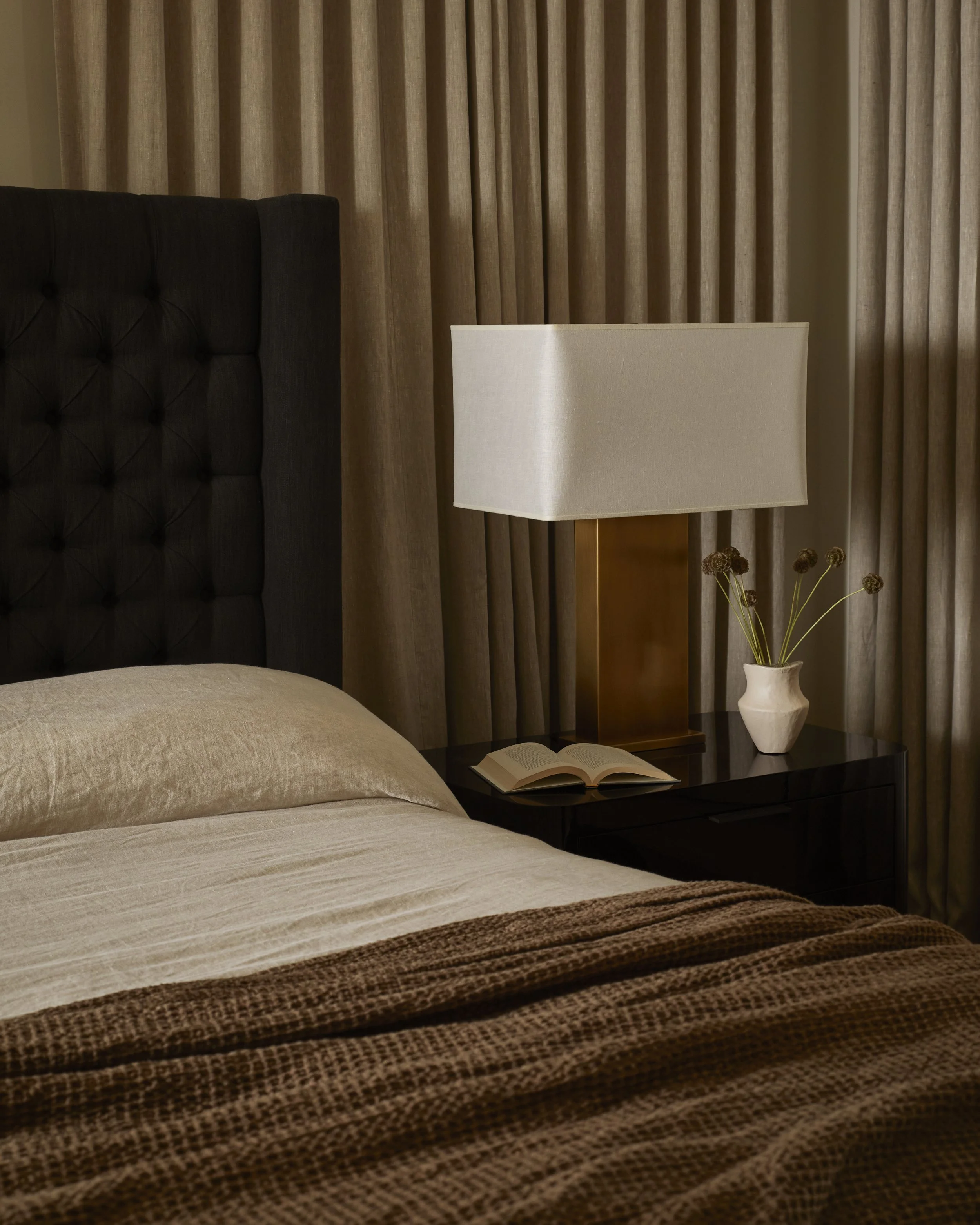 A cozy bedroom with beige bedding, a dark wooden bed frame, a bedside table with a white lampshade, a small open book, a decorative vase with dried flowers, and curtains in the background.