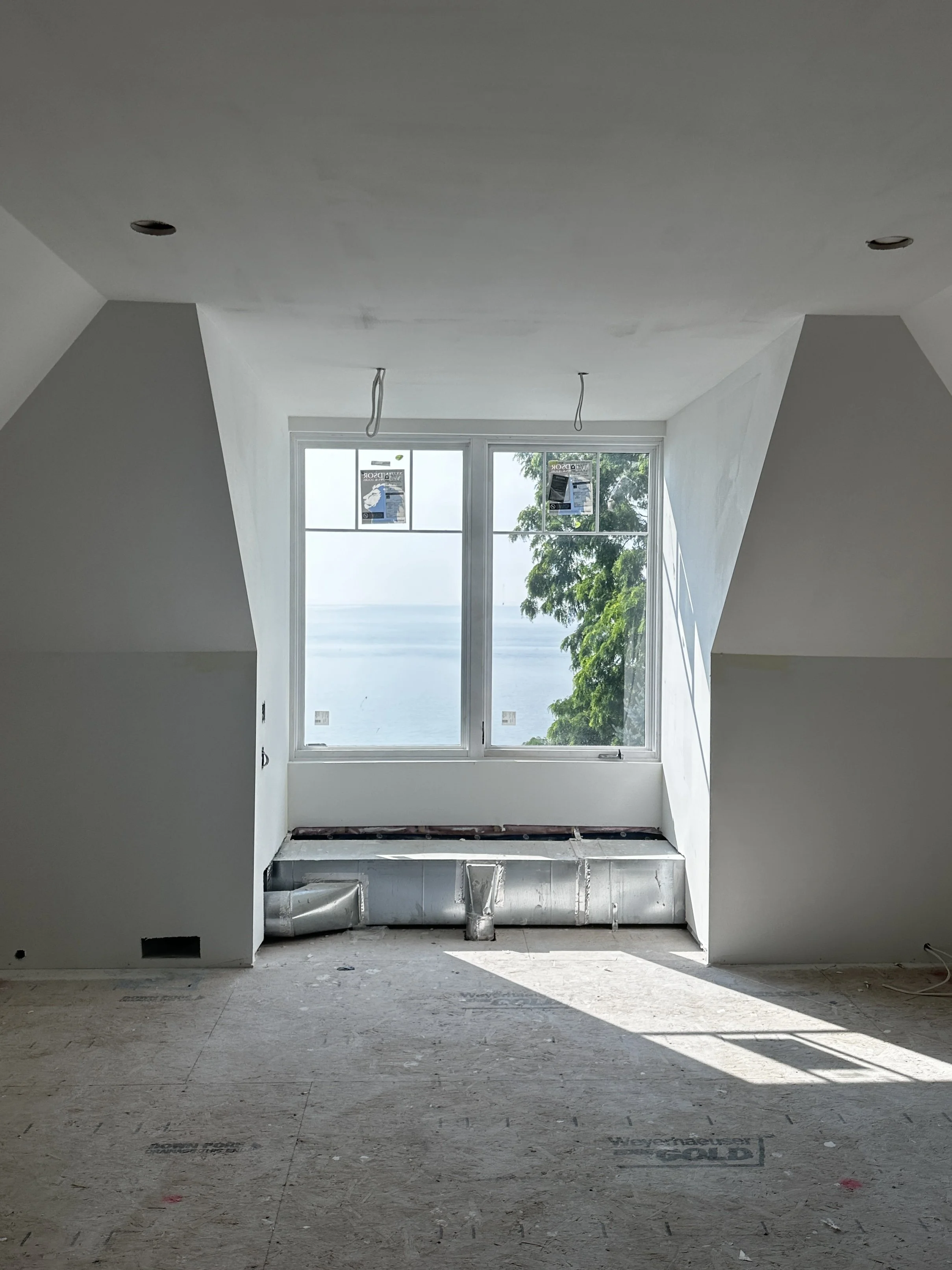Construction site of an unfinished room with a large window overlooking the ocean and trees, with drywall and ductwork installed.