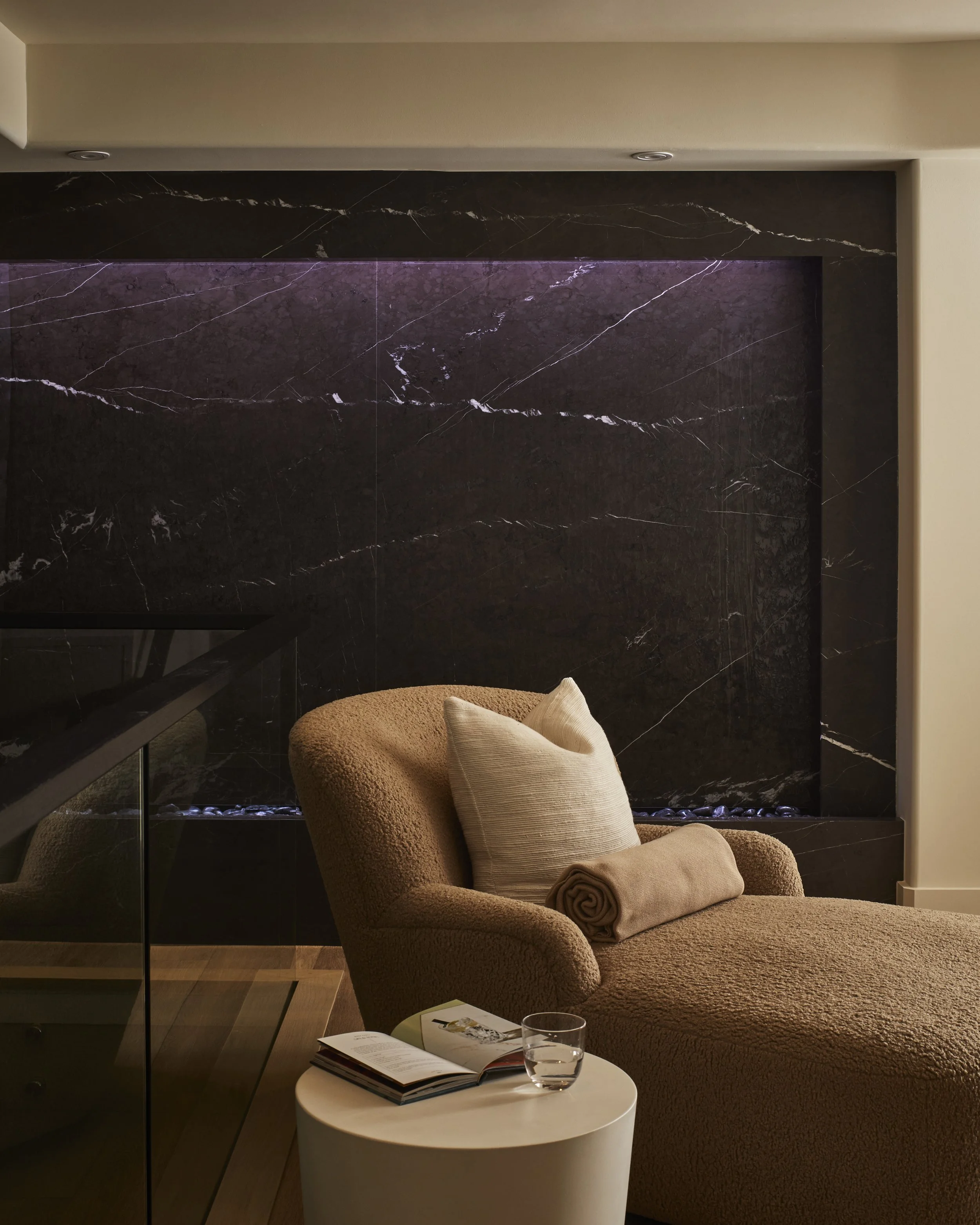 A cozy living room corner with a tan textured armchair and matching ottoman, a rolled blanket, a beige pillow, a white side table with an open magazine, a glass of water, a marble fireplace with dark stone and purple lighting, and a book on the table.