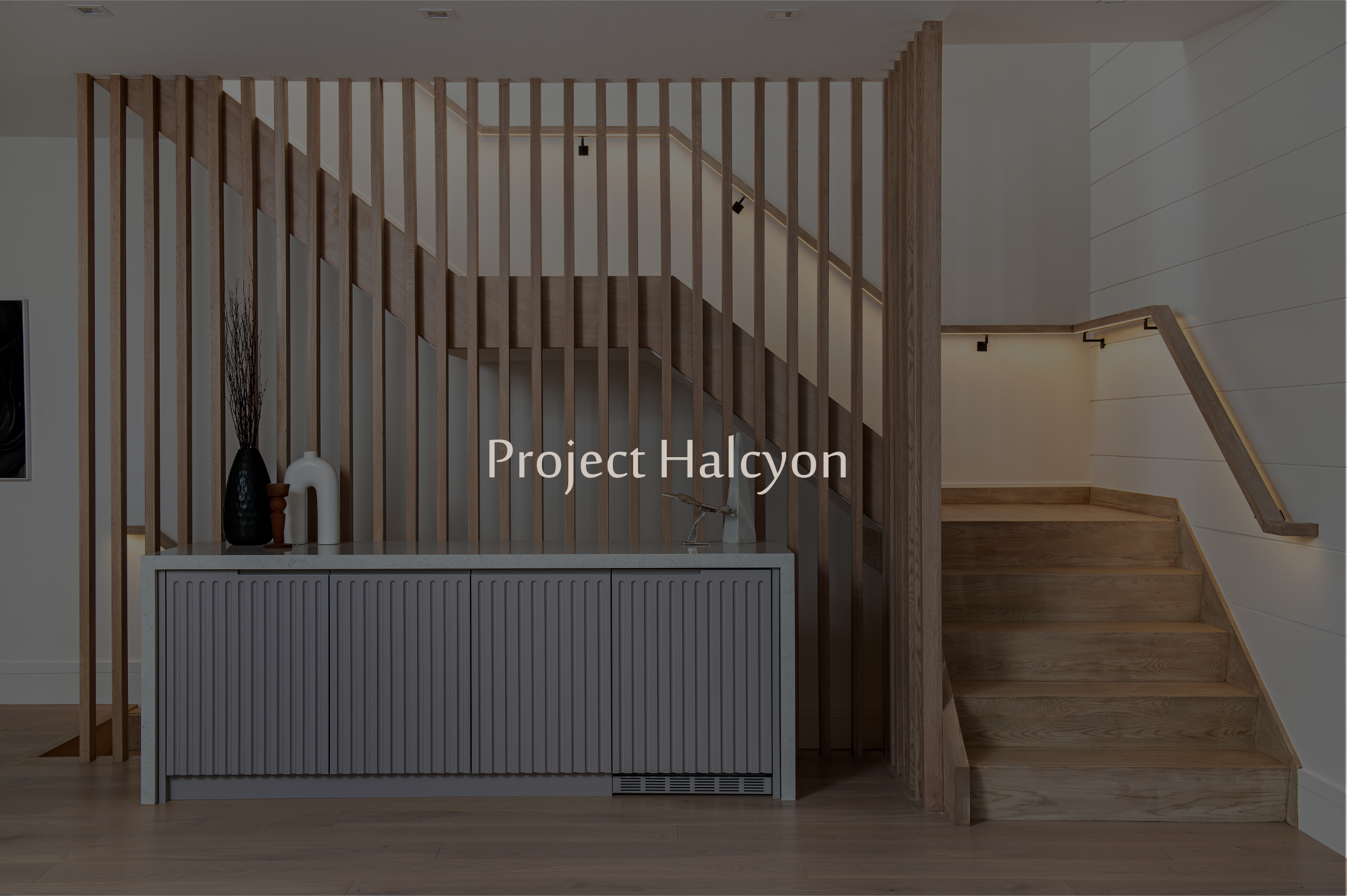 Interior view of a staircase with a wooden handrail and vertical wooden slats, a white cabinet with decorative vases on top, and a wall with horizontal wood paneling, illuminated by subtle lighting.