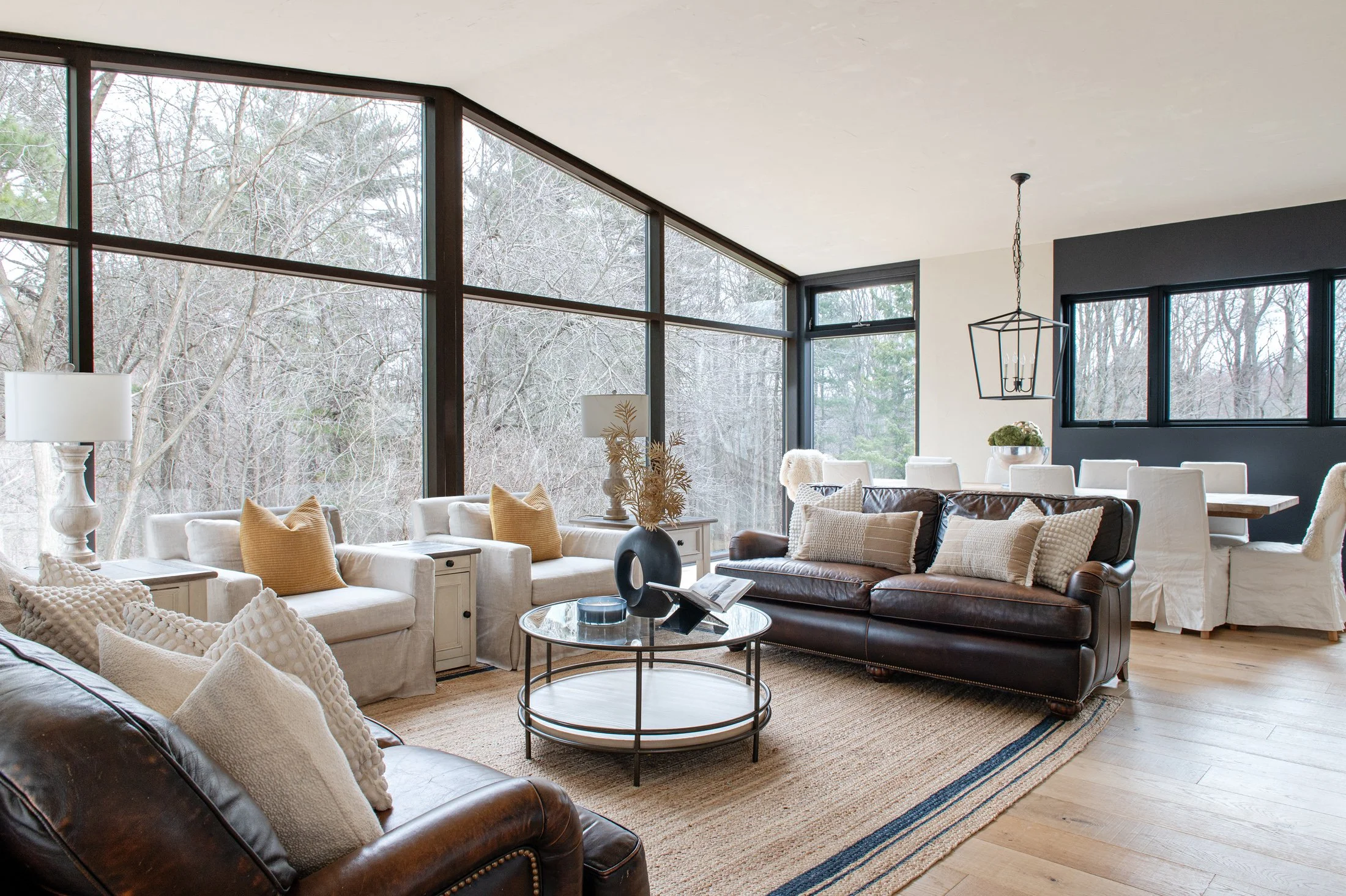 Living room with large glass windows showing trees outside, beige and brown furniture, including a brown leather sofa and white chairs, a round coffee table, and decorative lamps.
