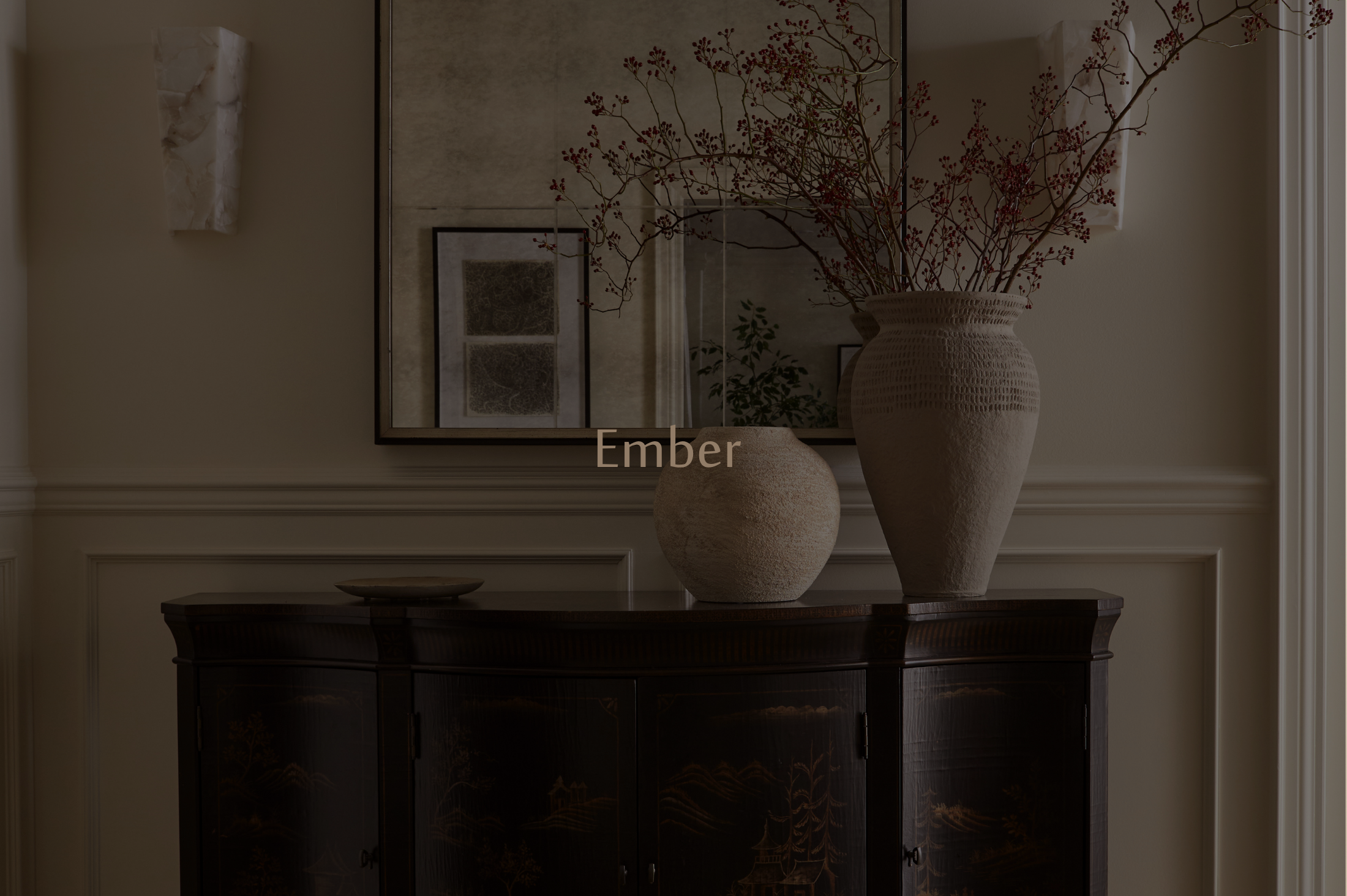 A beige wall decorated with framed artwork and a large vase with bare branches and red berries on a dark wooden sideboard.