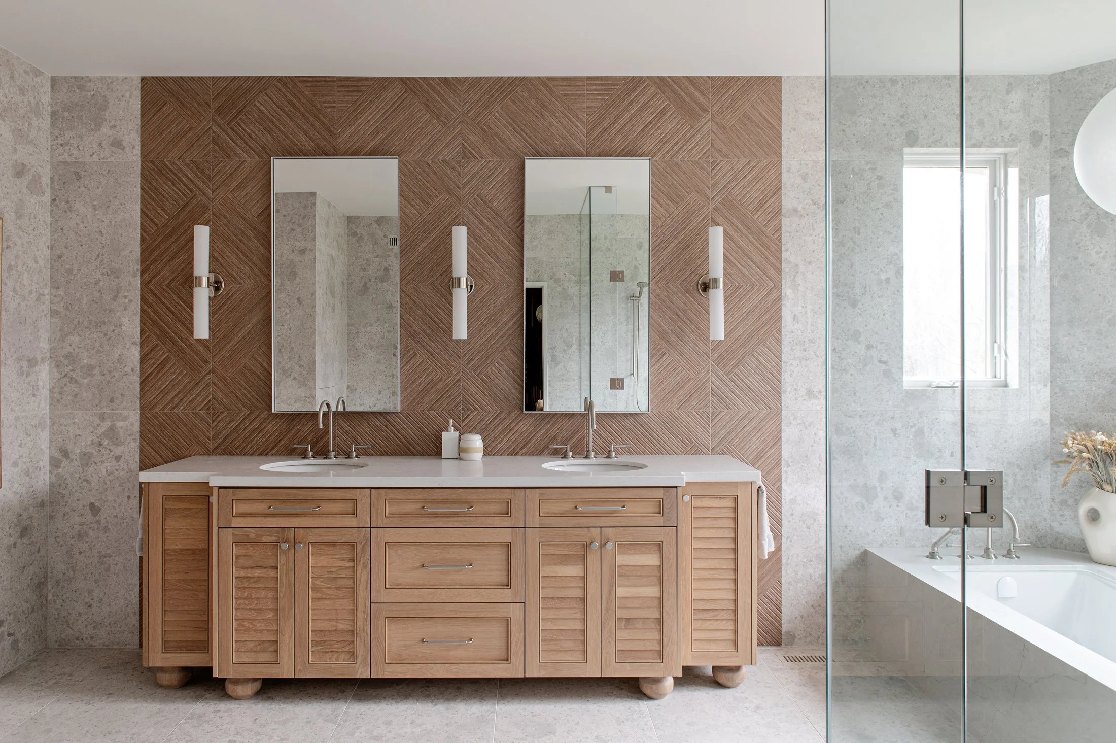 Modern bathroom with double vanity featuring a light wood cabinet, white countertop, and two mirrors. The wall behind the mirrors is decorated with textured, geometric wood panels. There are two slender wall-mounted light fixtures and a glass shower enclosure to the right, with a window providing natural light.