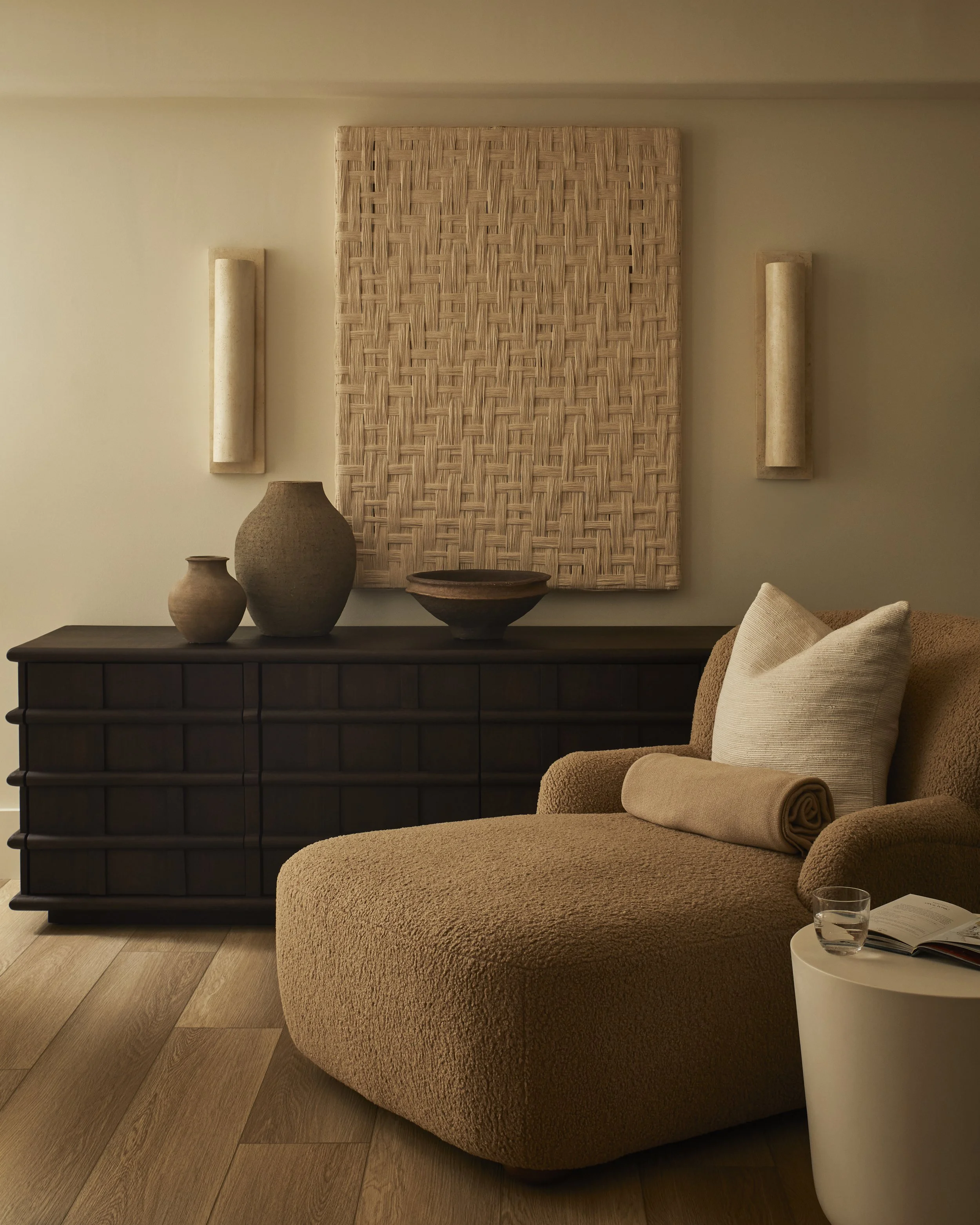 A beige chaise lounge with a rolled pillow, a dark wooden sideboard with three vases on top, dark wall art with geometric patterns, and a beige cylindrical wall lamps on a white wall, with a wooden floor.
