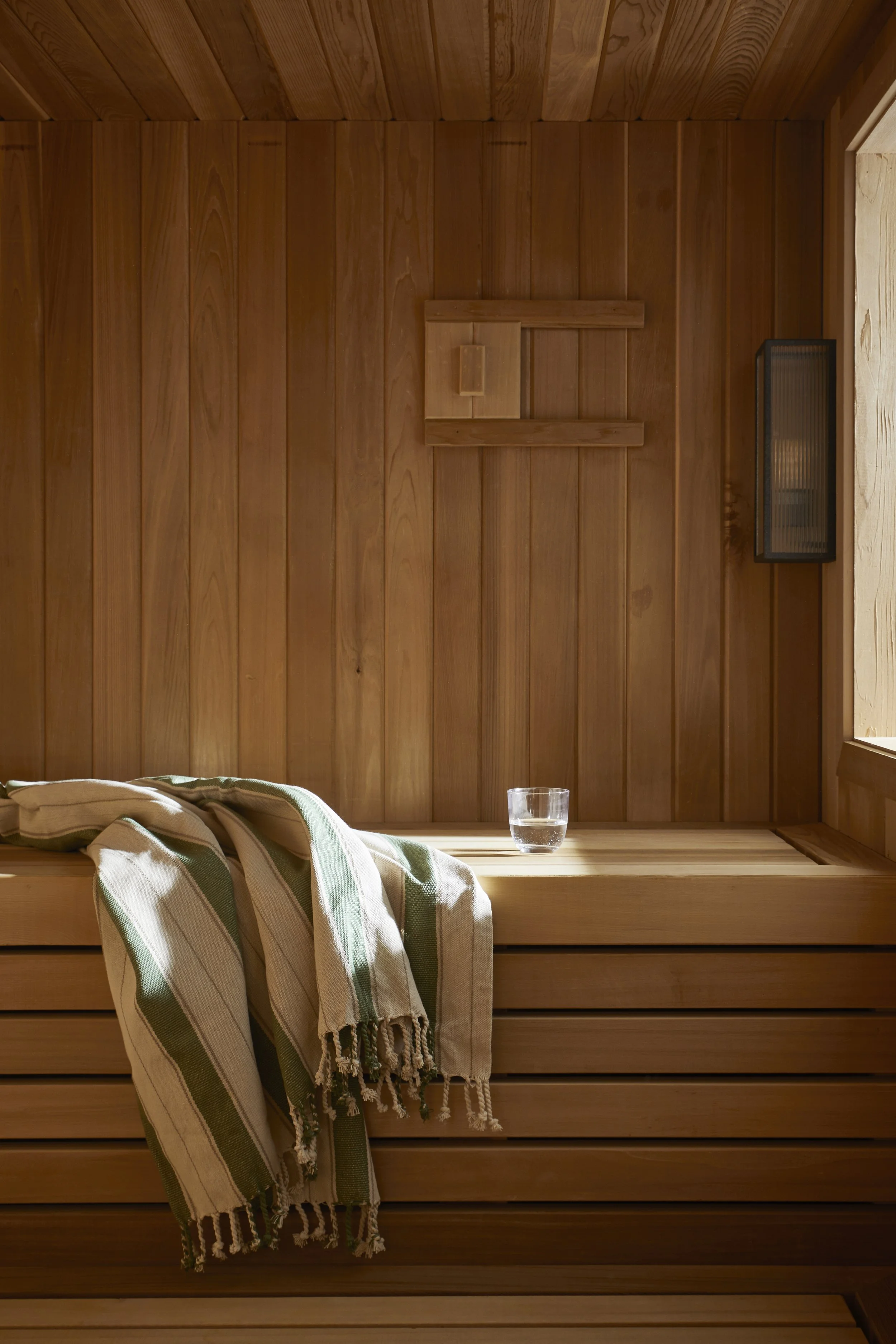 A wooden sauna interior with vertical wood paneling, a small wooden shelf, a black wall-mounted heater, a glass of water on a wooden ledge, and a striped towel draped over the edge of the shelf.