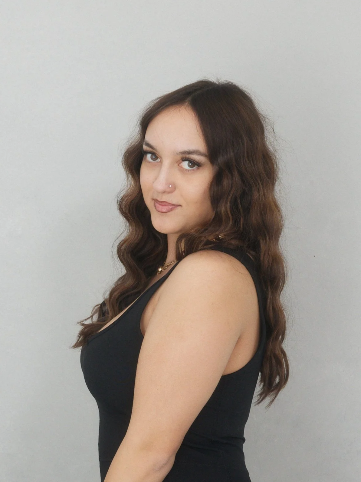 A young woman with long, wavy brown hair and light skin posing against a plain light-colored wall. She is wearing a black sleeveless top and a gold necklace, looking at the camera with a slight smile.