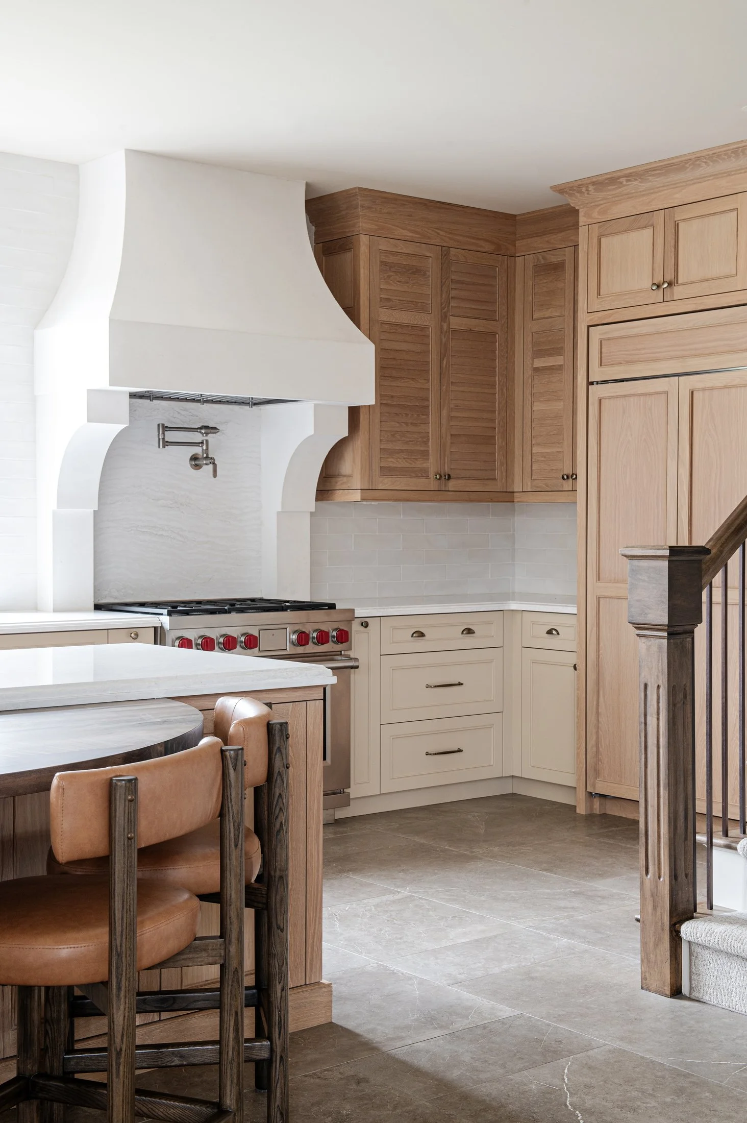 Kitchen with white range hood, wood cabinets, cream lower cabinets, beige flooring, and a dining table with brown leather chairs.