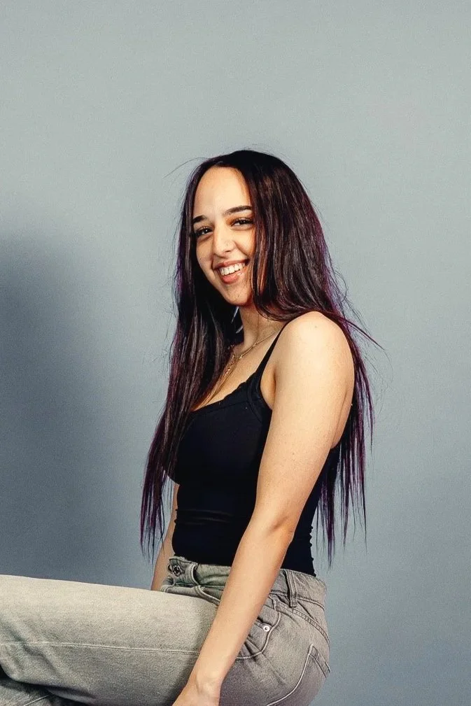A young woman with long dark hair and purple highlights, smiling and winking at the camera, wearing a black tank top and light gray jeans, sitting against a plain gray background.