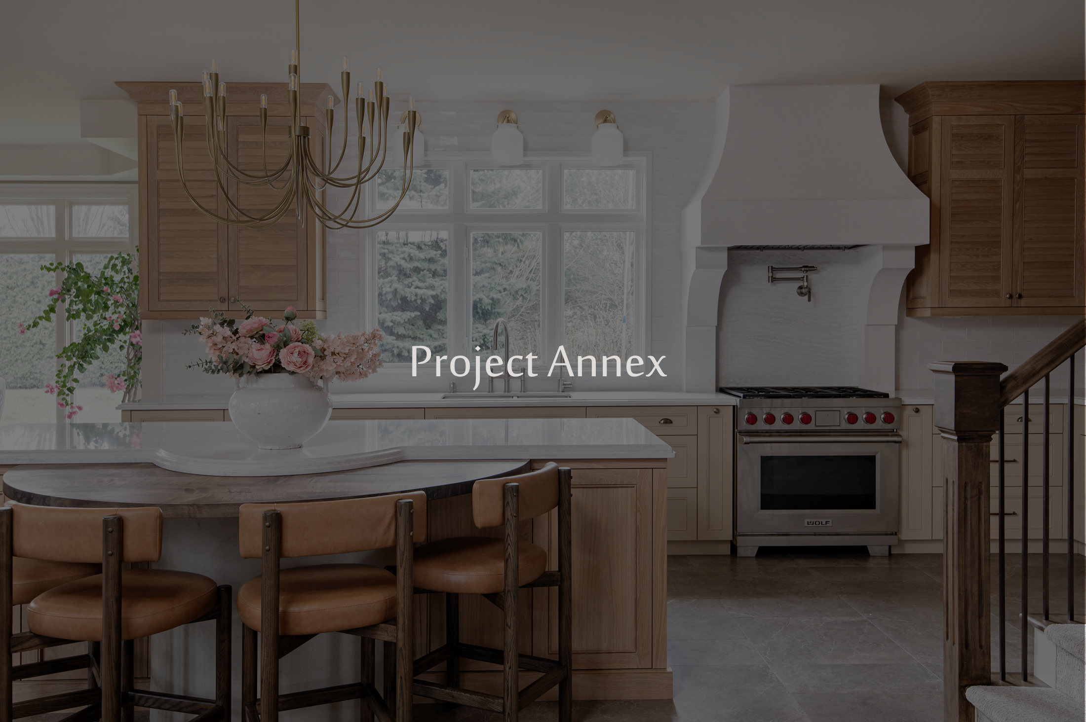 A kitchen with wooden cabinets, a white fireplace, large window, a round dining table with wooden chairs, a chandelier, and a vase with pink flowers.