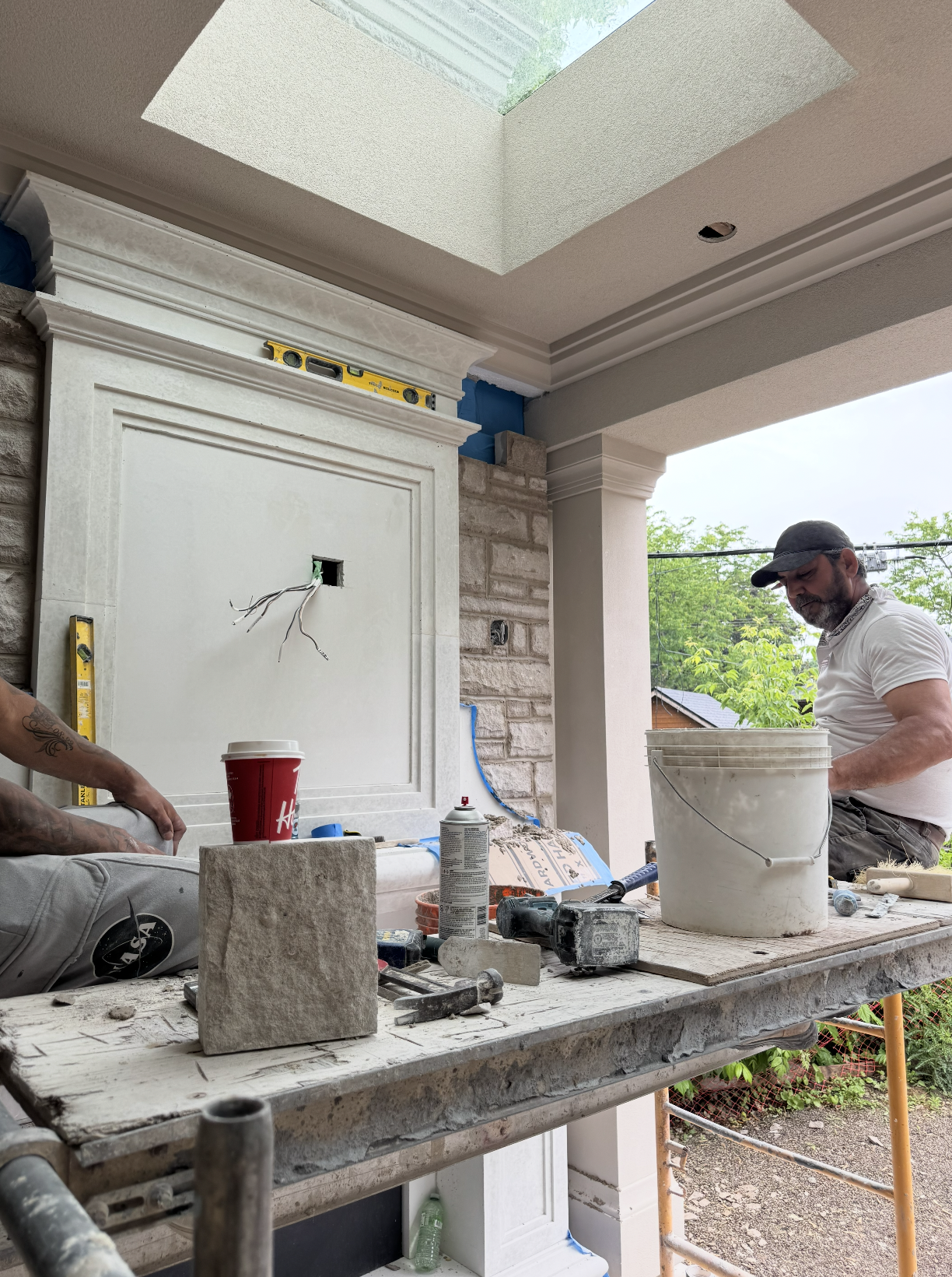 A man sits on a construction site working on a building or renovation project outdoors, with tools, materials, and equipment on a worktable in front of him. The background features part of a house's exterior wall and trees outside.