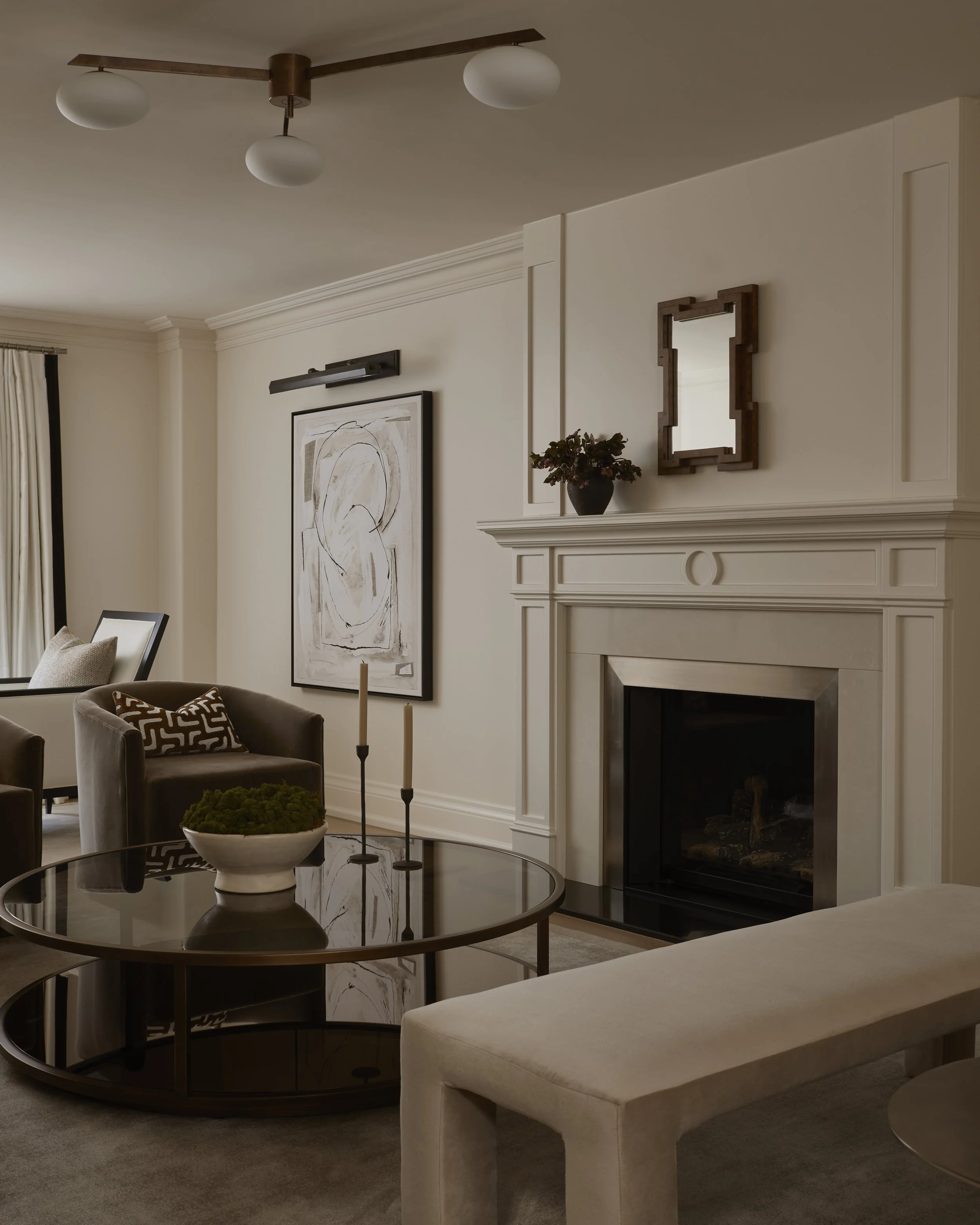 Living room with contemporary decor, including a black wooden fireplace, a framed abstract art piece, a round glass coffee table with a decorative bowl, a beige upholstered chair, a black framed mirror, and a dark ceiling light fixture with three globe bulbs.