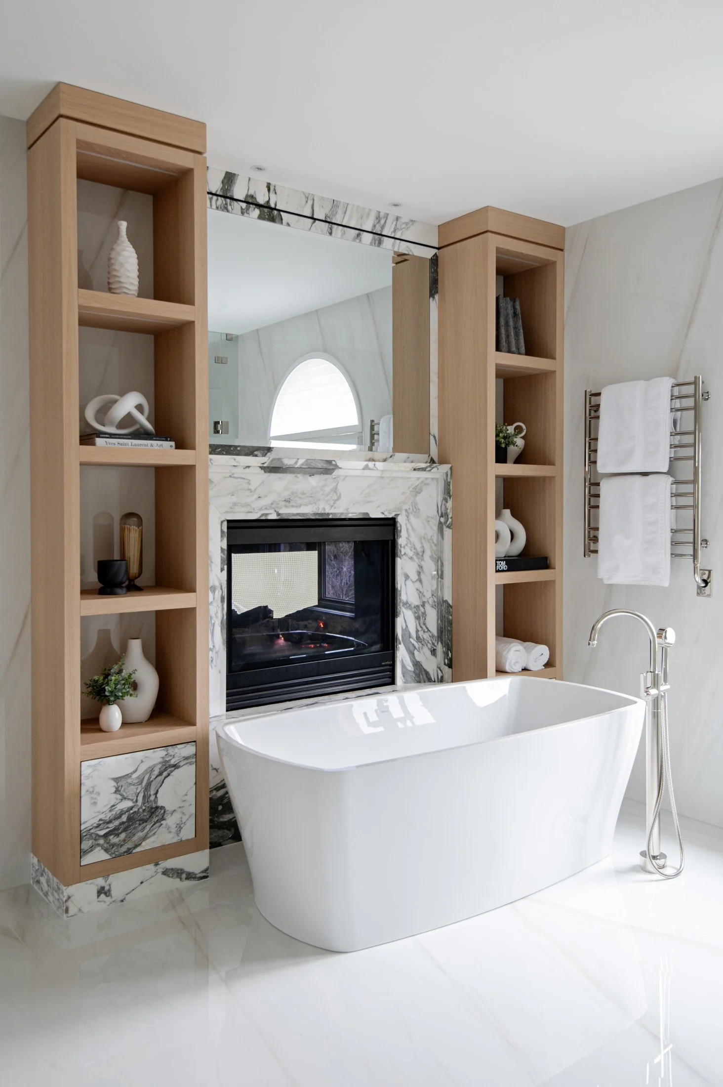 Modern bathroom with a freestanding white bathtub, marble fireplace with a mirror above, and open wooden shelves with decorative objects, towels, and plants.