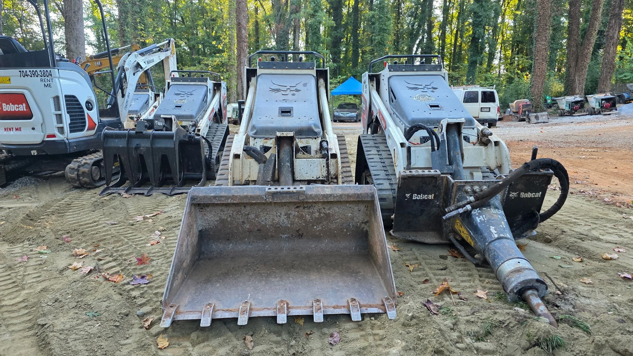 Construction equipment including three small skid steer loaders and a compact excavator parked on a dirt surface with trees and parked vehicles in the background.