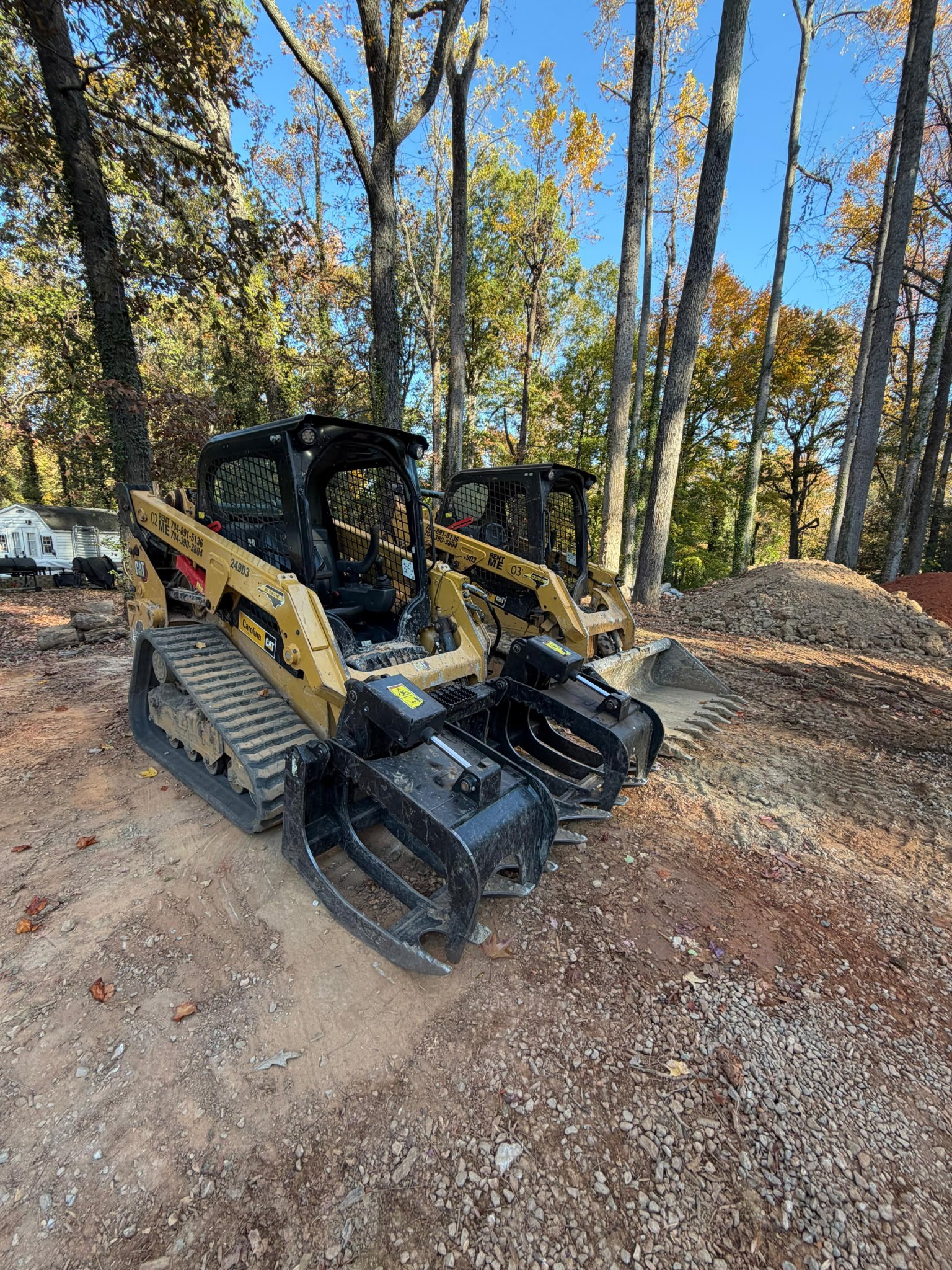 Two compact track loaders parked on a dirt surface with trees in the background.