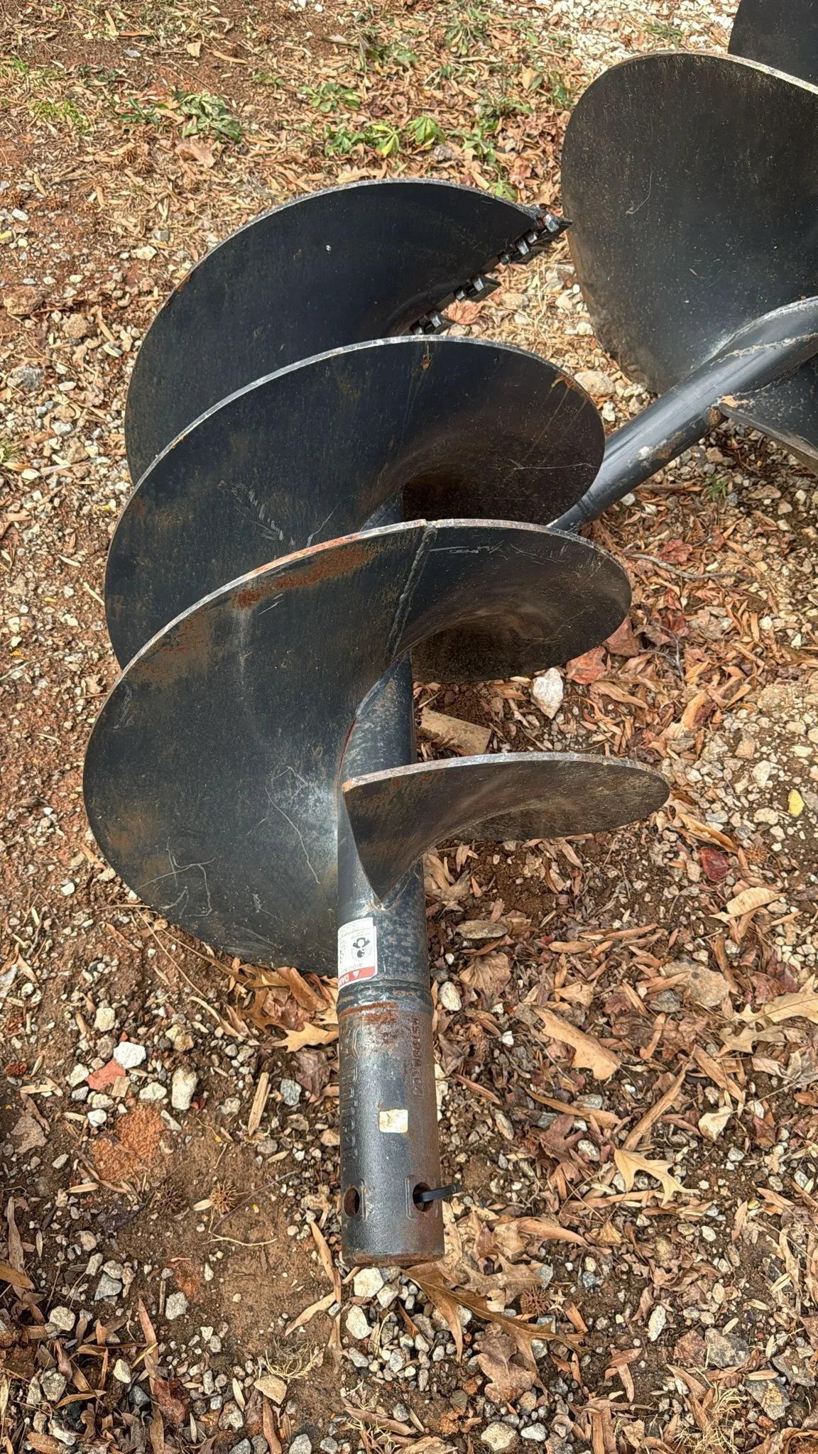 A metal auger drill bit lying on dirt ground with small rocks and fallen leaves.