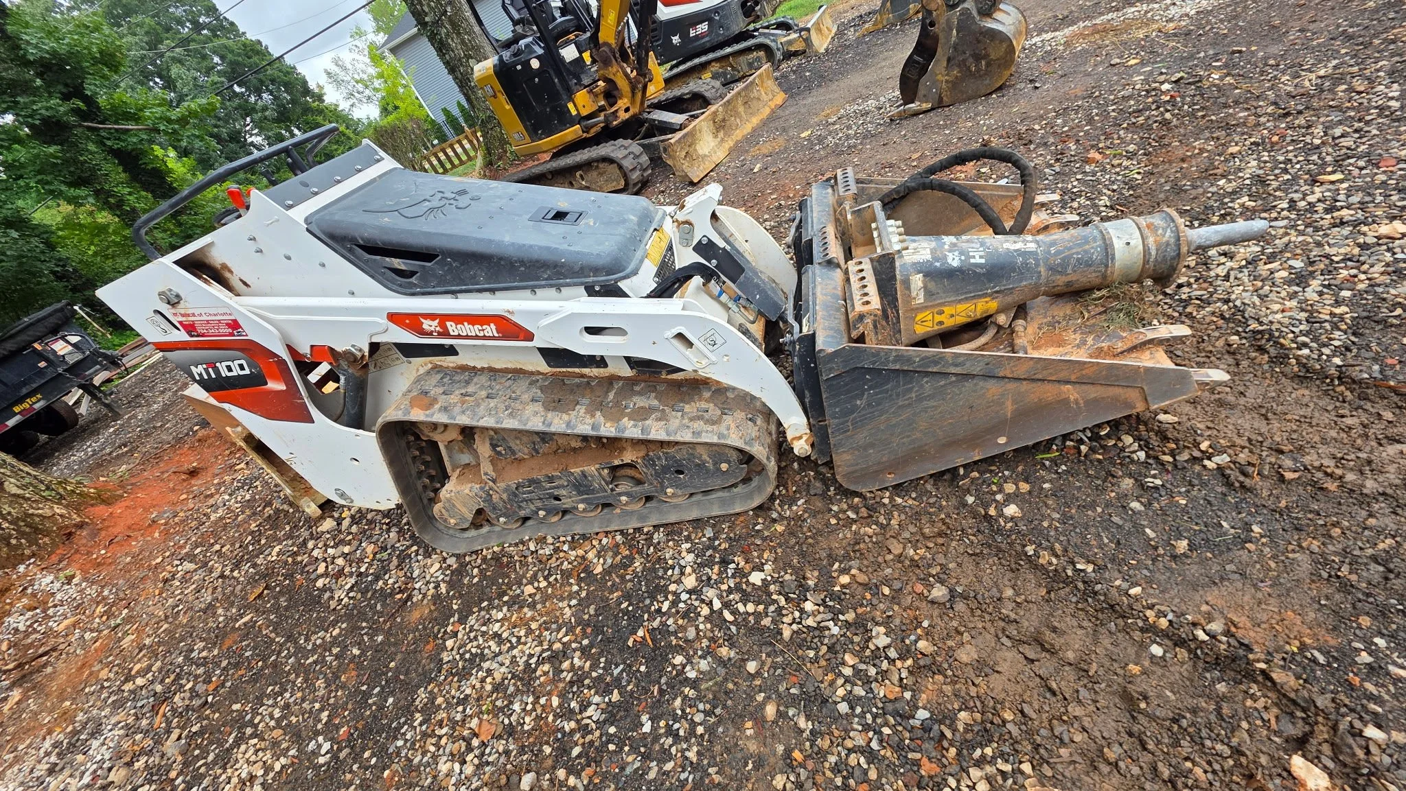 A tracked Bobcat compact track loader with a backhoe attachment resting on rocky ground outdoors, with trees and other construction equipment in the background.