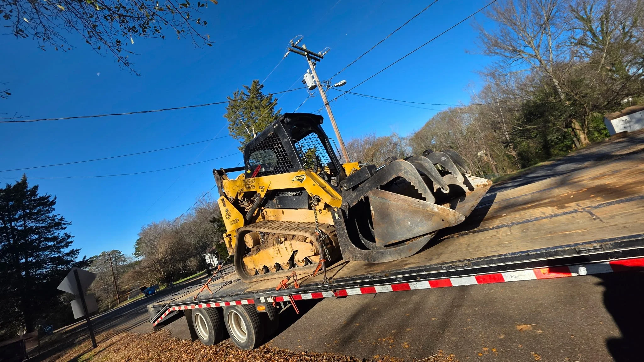 A yellow bulldozer on a flatbed trailer being transported on a road, with clear blue sky and trees in the background.