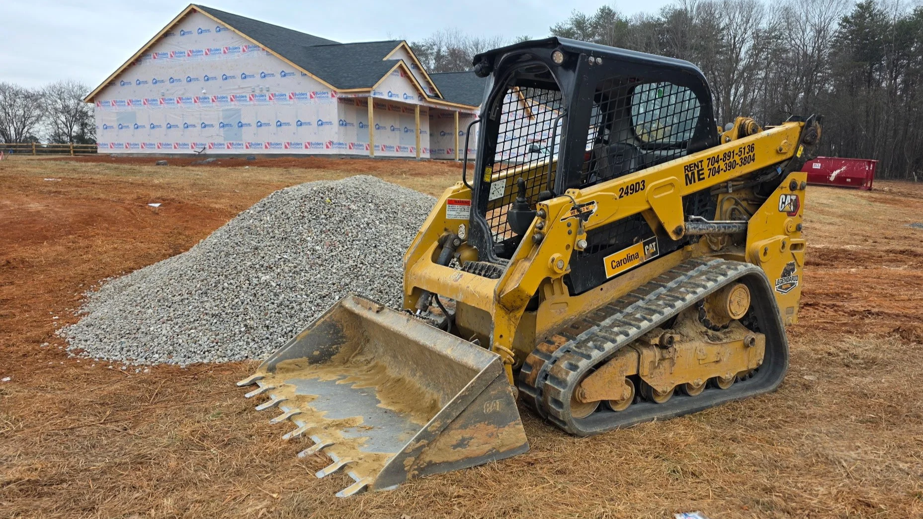 Construction site with a small yellow Caterpillar skid steer loader on dirt ground, a pile of gravel nearby, and a house under construction in the background.
