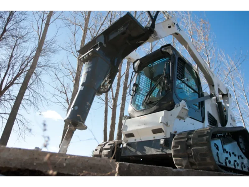 A white and black mini excavator with a hydraulic arm and bucket attachment, working outdoors among leafless trees.