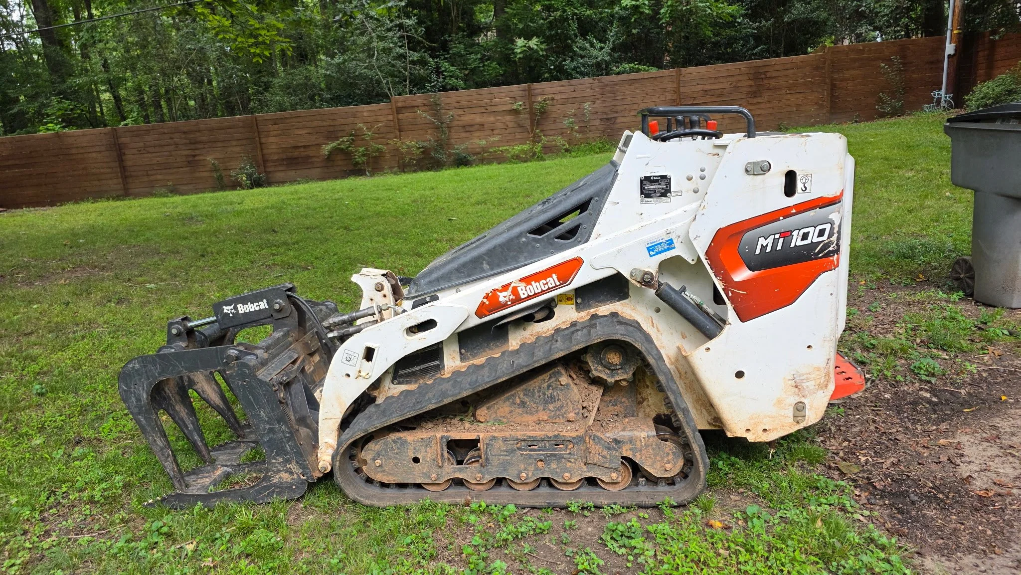 A Bobcat MT 100 compact track loader with a black grapple attachment, parked on a grassy area next to a wooden fence and a trash bin, with trees in the background.