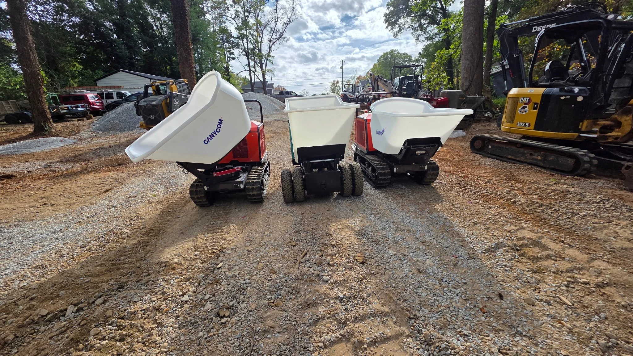 Three small remote-controlled dumper trucks on a construction site with trees and construction equipment in the background.