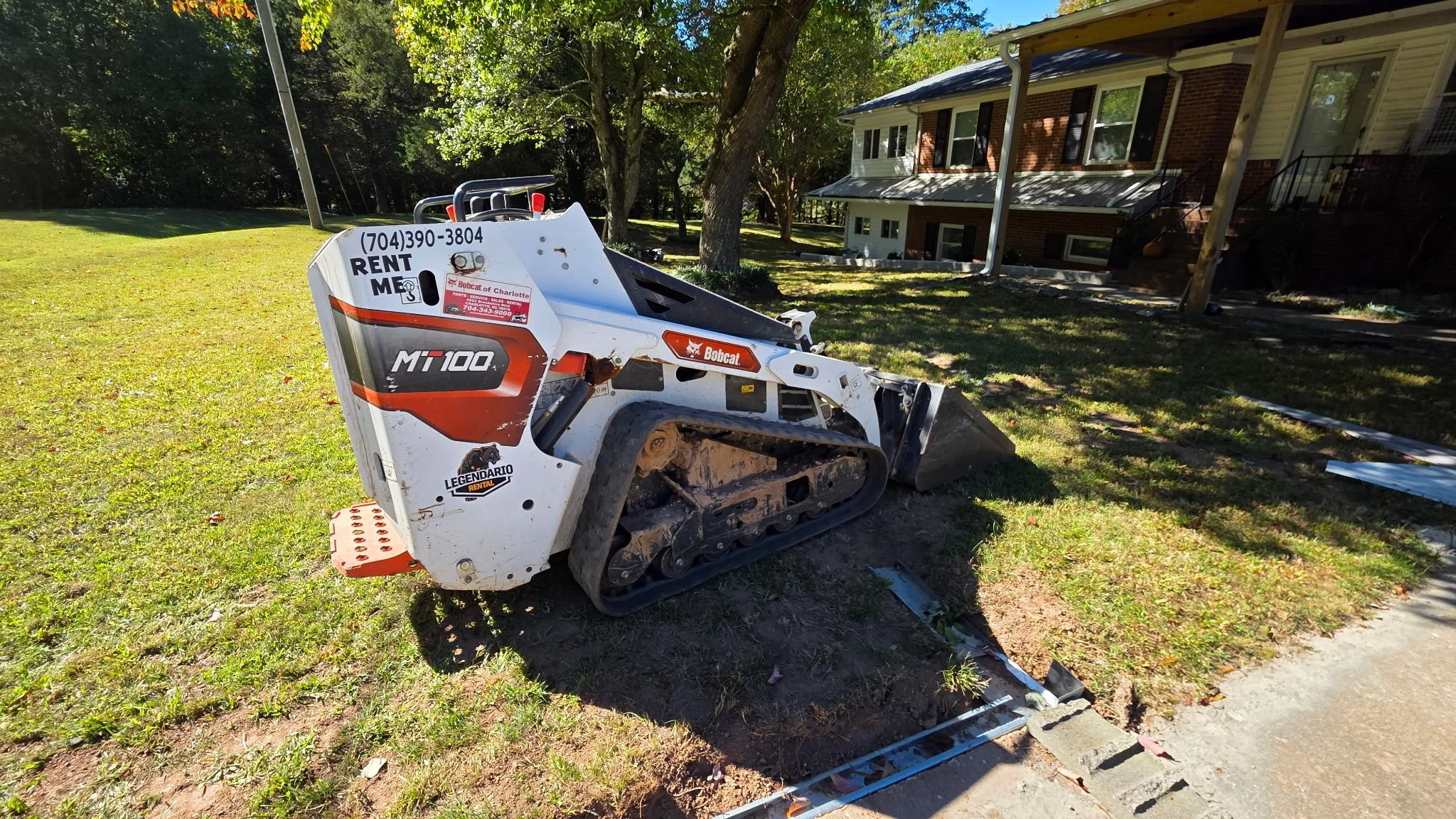 A small white Bobcat MT100 compact loader with tracks, parked on a grassy area near a residential house, with construction materials scattered nearby.