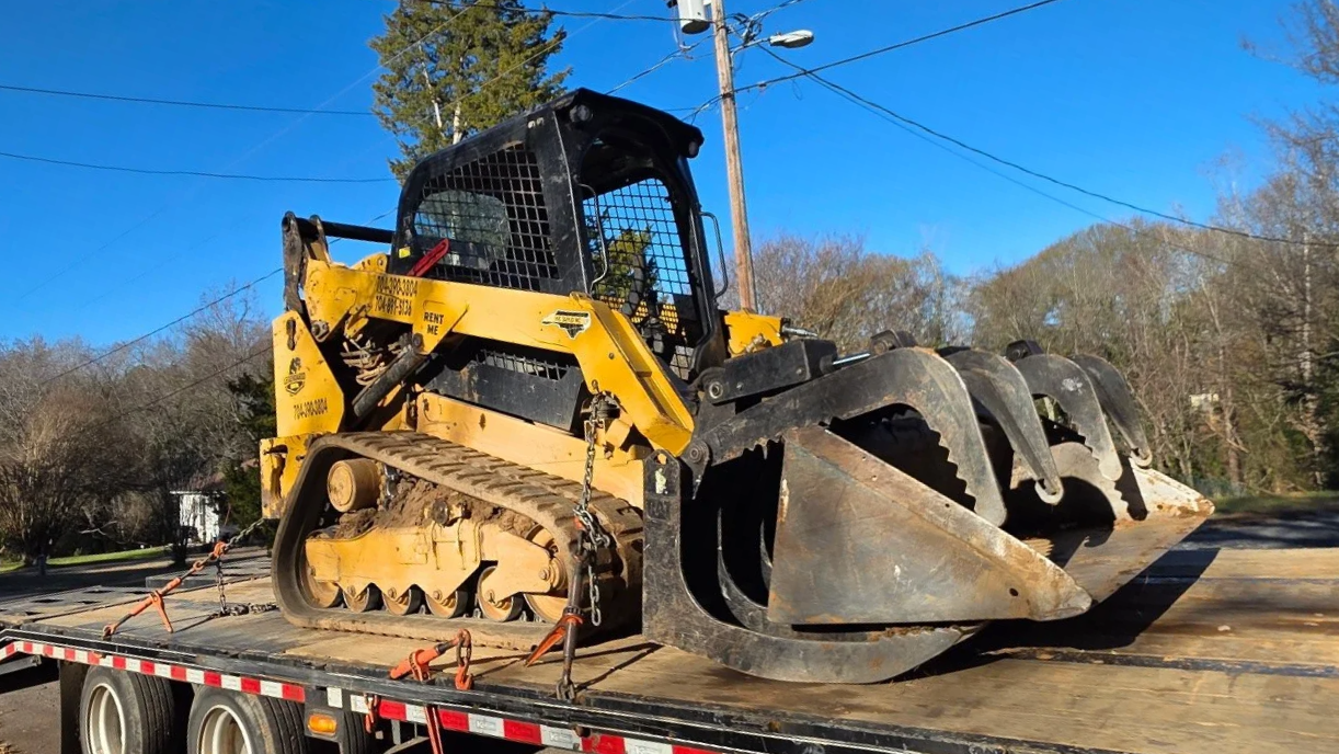 A large yellow and black bulldozer loaded on a flatbed truck, secured with chains, on a road with trees and power lines in the background.