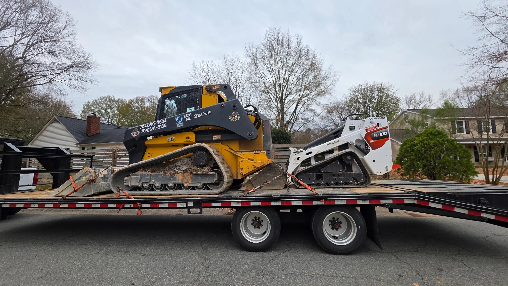A flatbed truck carrying two compact tracked utility machines, one yellow and one white, on a residential street with houses and trees in the background.