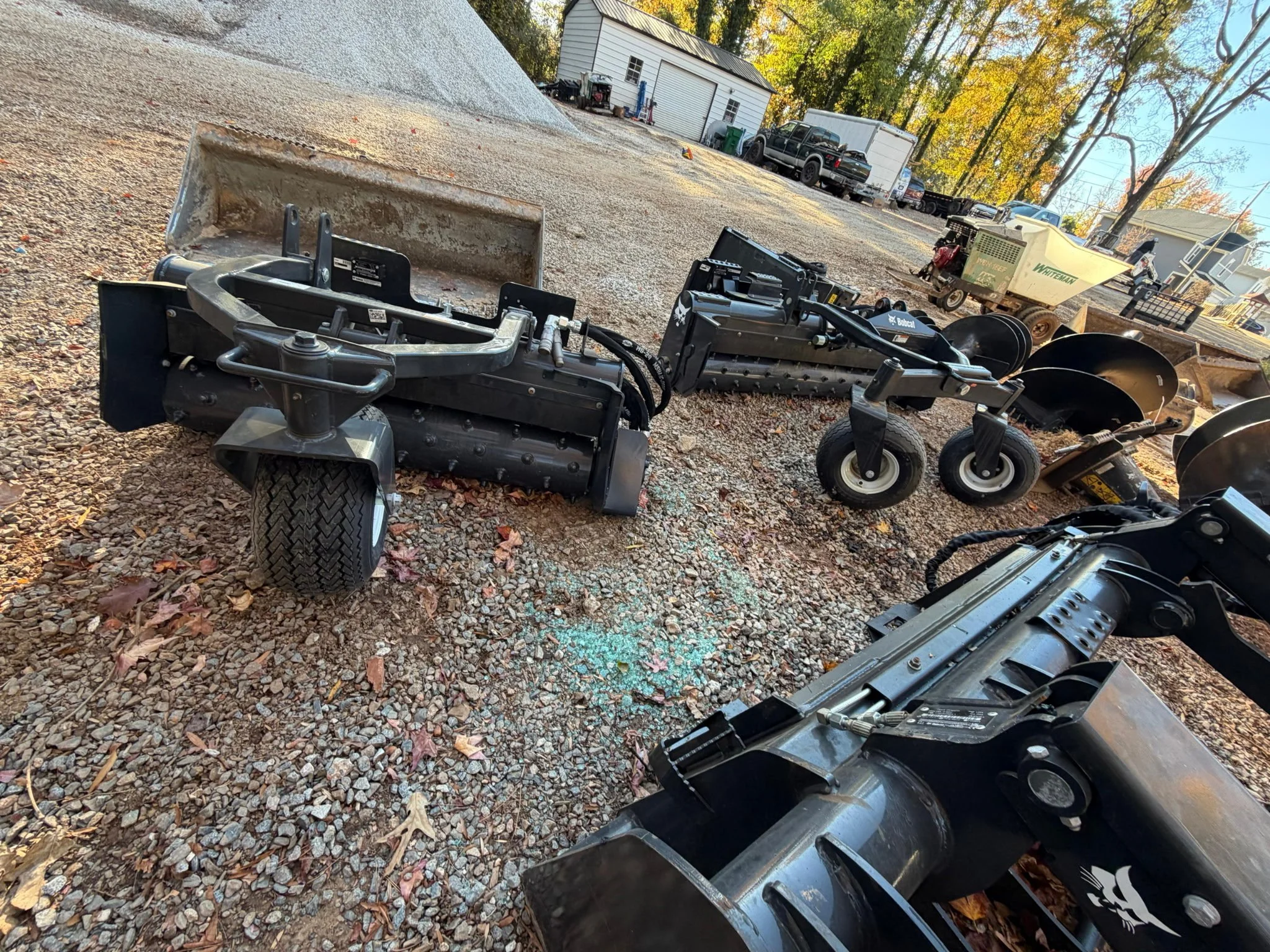 Various pieces of construction or landscaping equipment and attachments, including a large plow, a tow-behind roller, and a small tracked vehicle, are arranged outdoors on a gravel surface with trees and houses in the background.