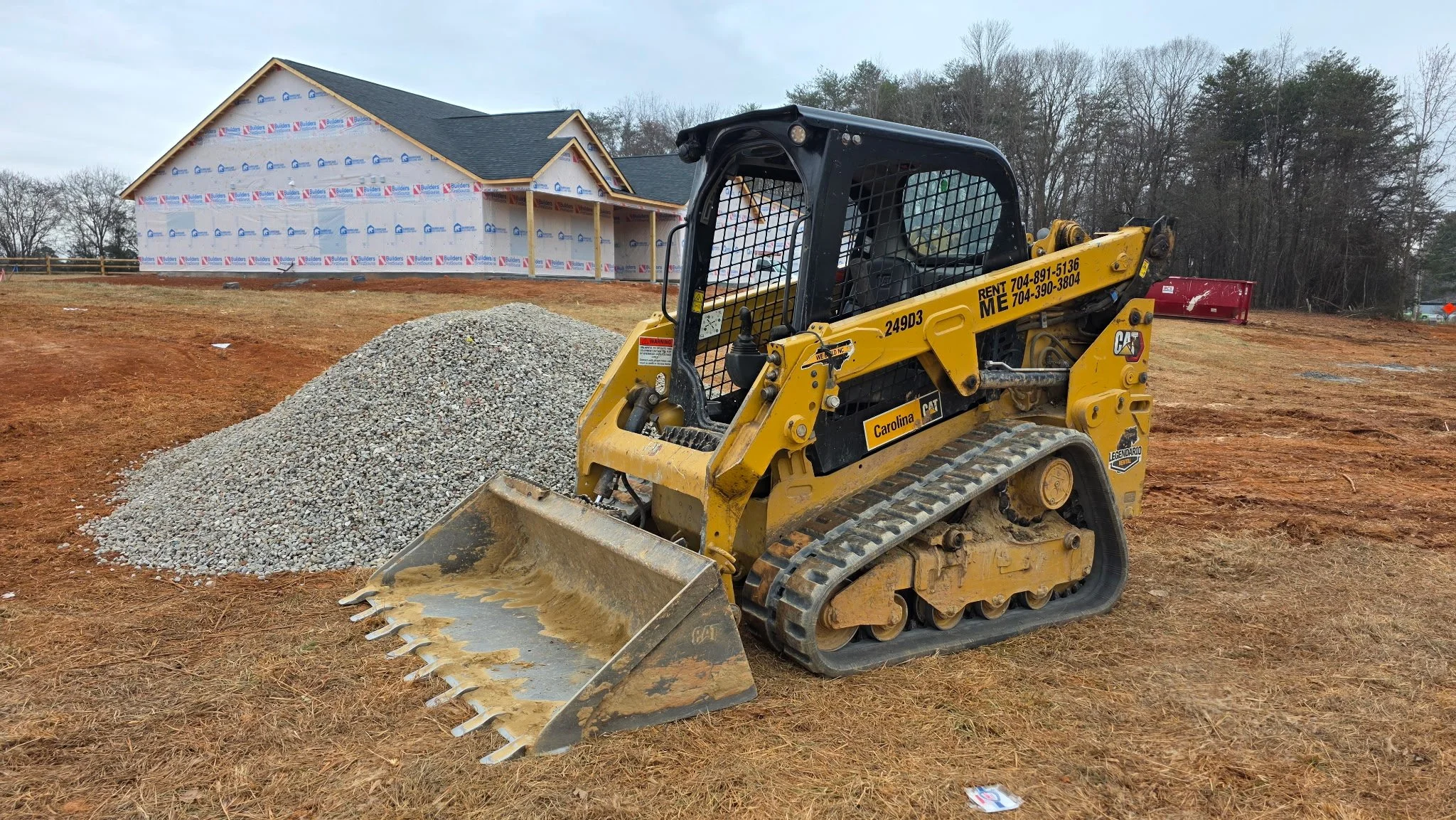 Construction site with a yellow CAT mini track loader and a pile of gravel in front of a house under construction.