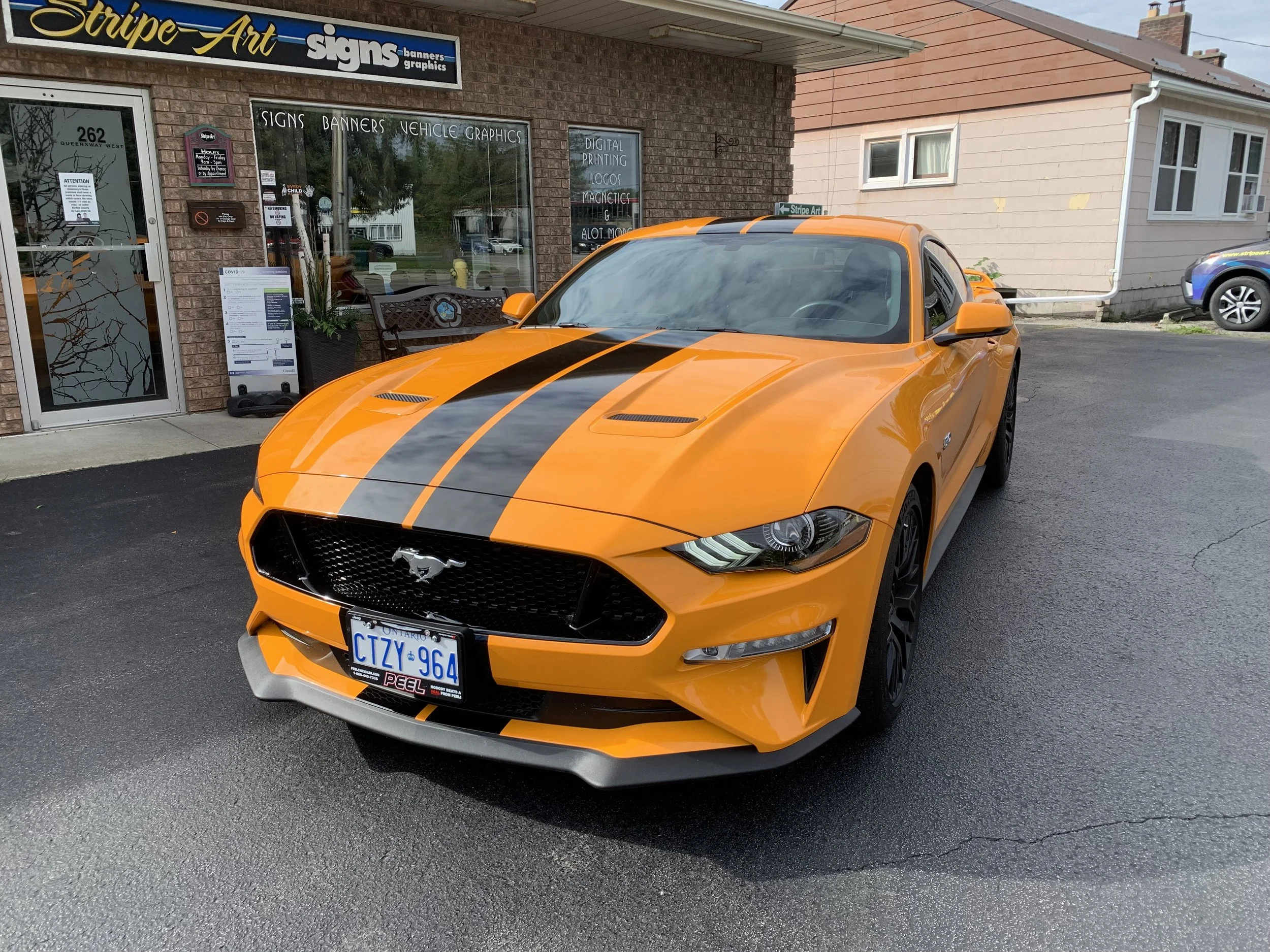 Orange Ford Mustang with black racing stripes parked outside a sign shop.