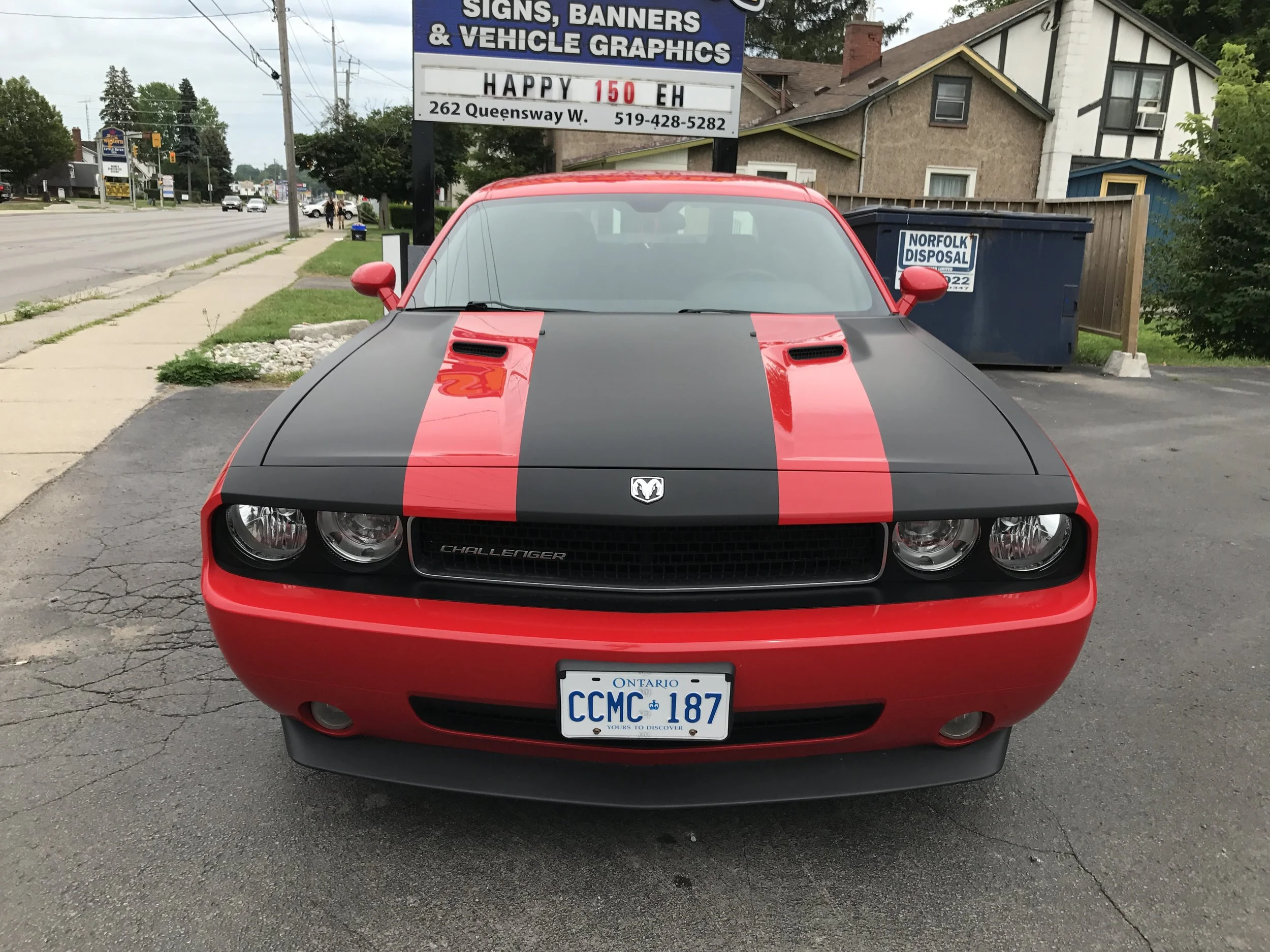Front view of a red and black Dodge Challenger car parked on a street. The car has a black hood with two red racing stripes. The vehicle displays an Ontario license plate reading 'CCMC 187'. In the background, there is a sign advertising signs, banners, and vehicle graphics, and a blue Norfolk Disposal dumpster.