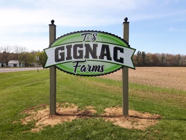 Sign for T&S Gignac Farms on a grassy field, with a dirt patch beneath the sign, and open farmland in the background.