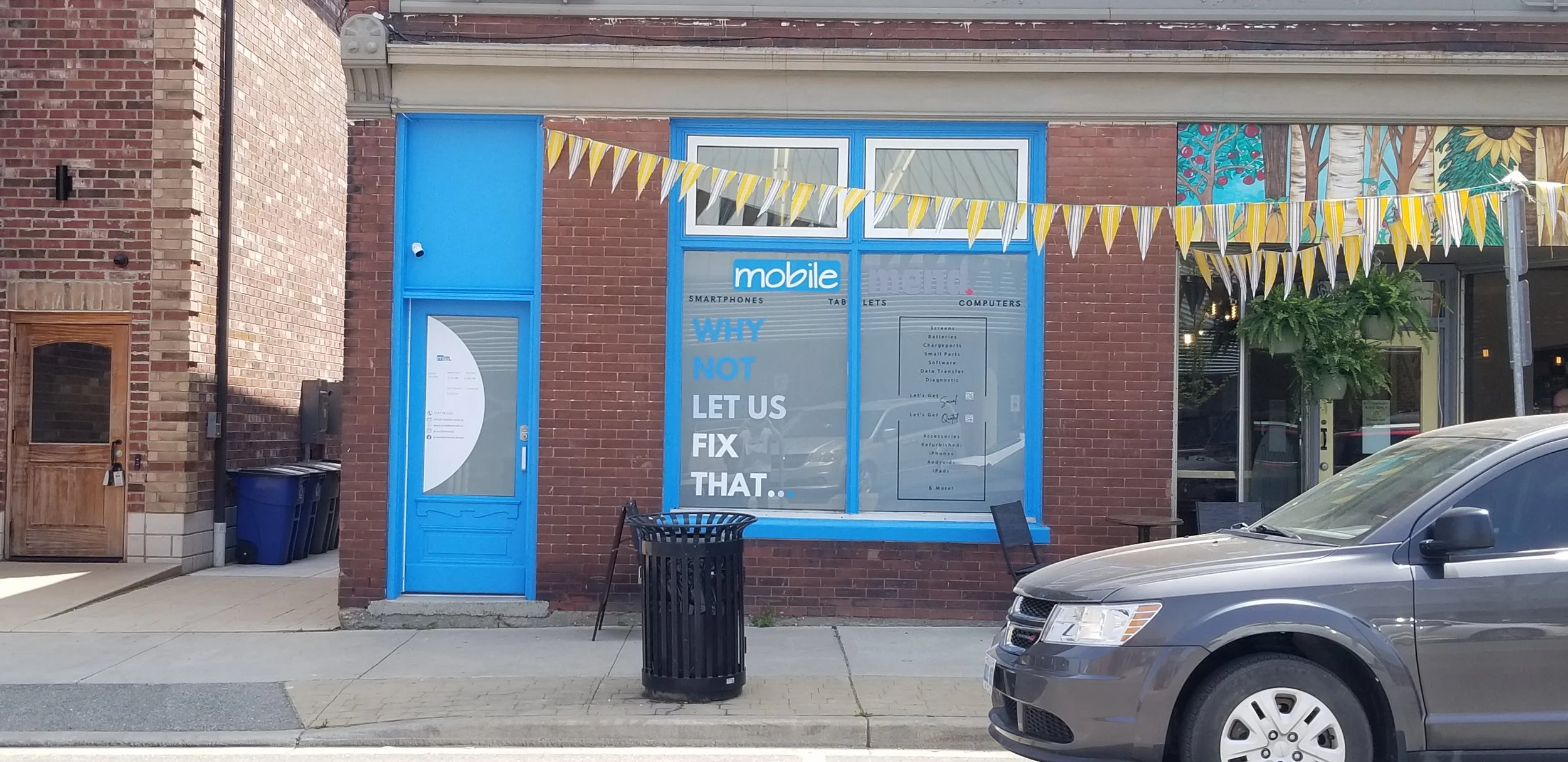 storefront with a blue door and window, yellow and white pennant banner, a black trash can, a parked gray vehicle, and mural art on the building wall.