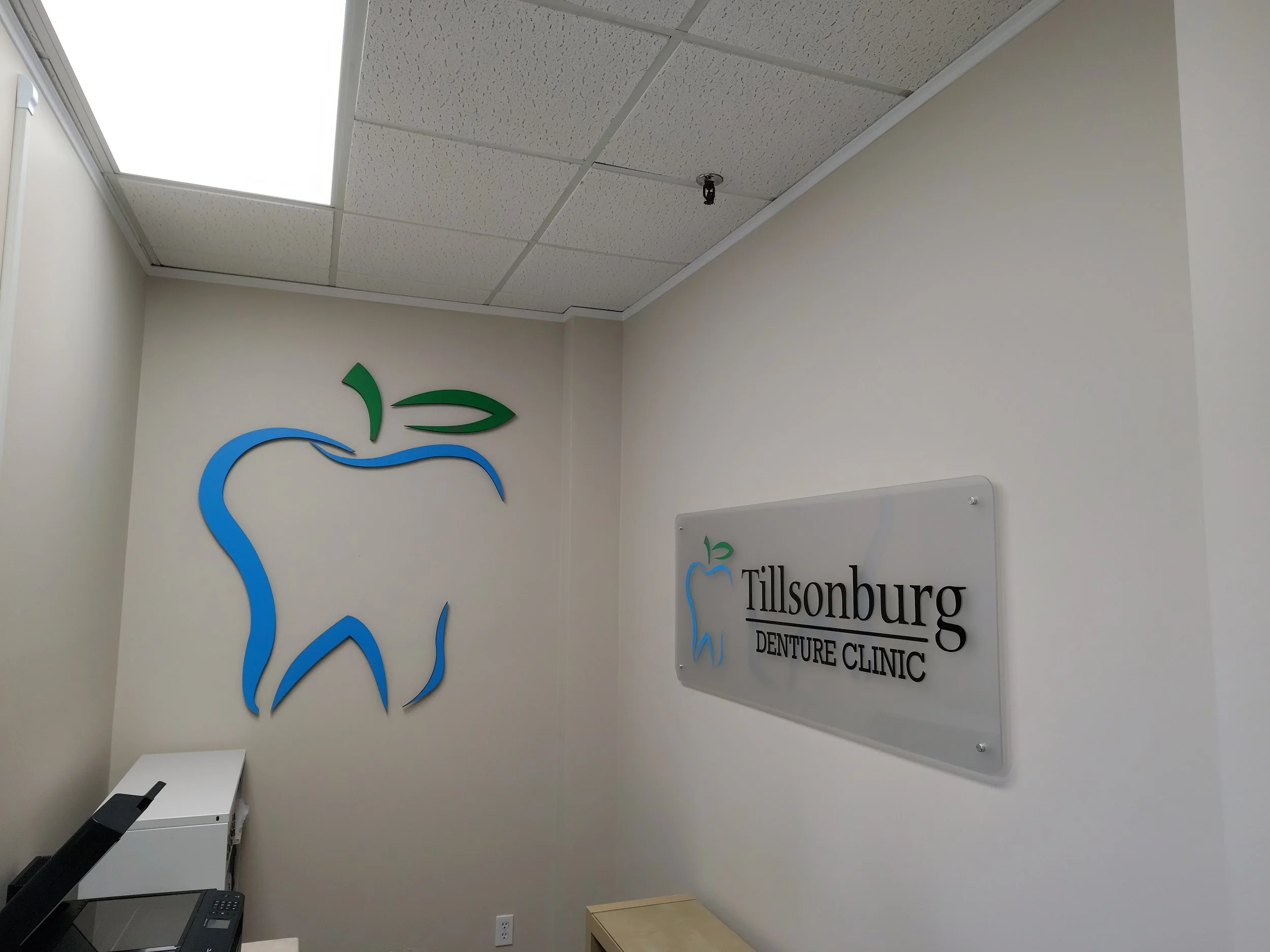 Interior of Tillsonburg Denture Clinic with a wall logo of a tooth outline with a green leaf and blue line art, and a matching sign on the wall.