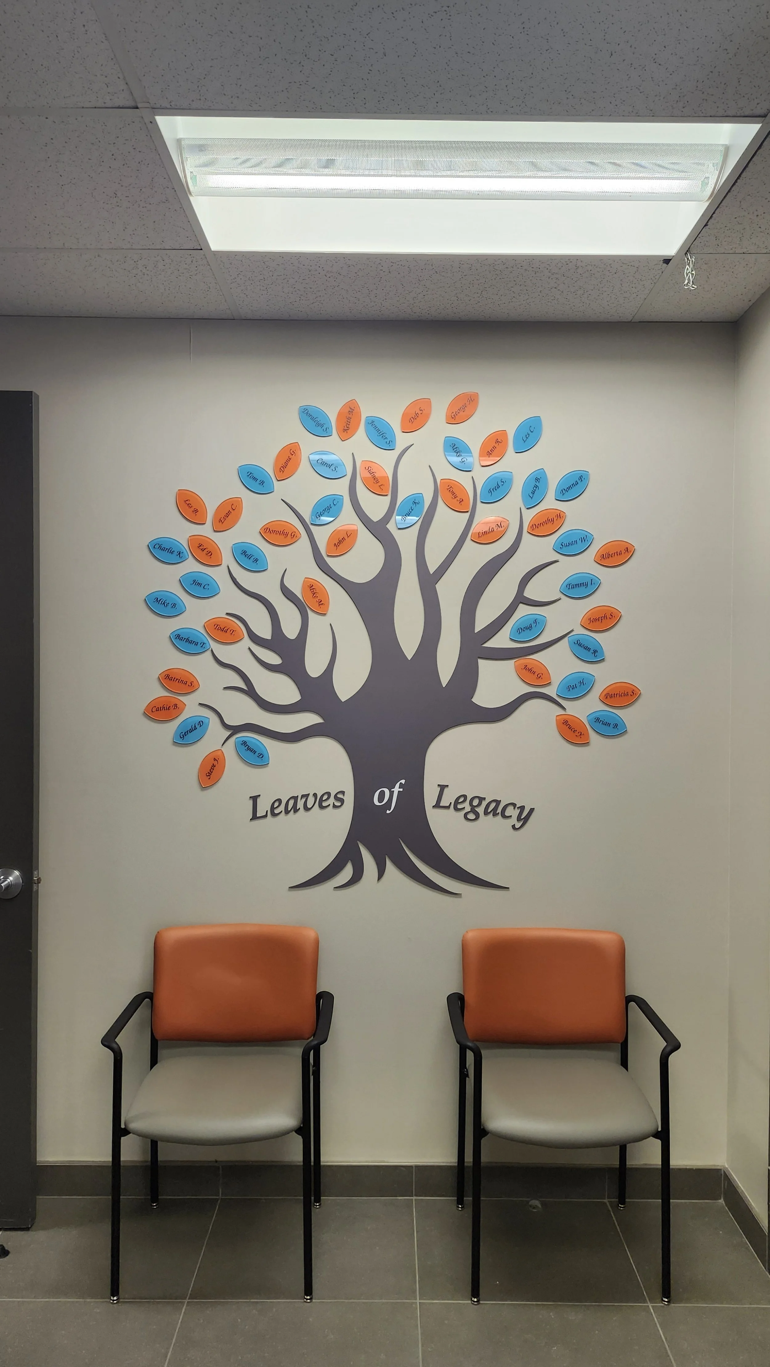Wall display of a tree with orange and blue name tags representing leaves, titled 'Leaves of Legacy,' with two chairs in front.