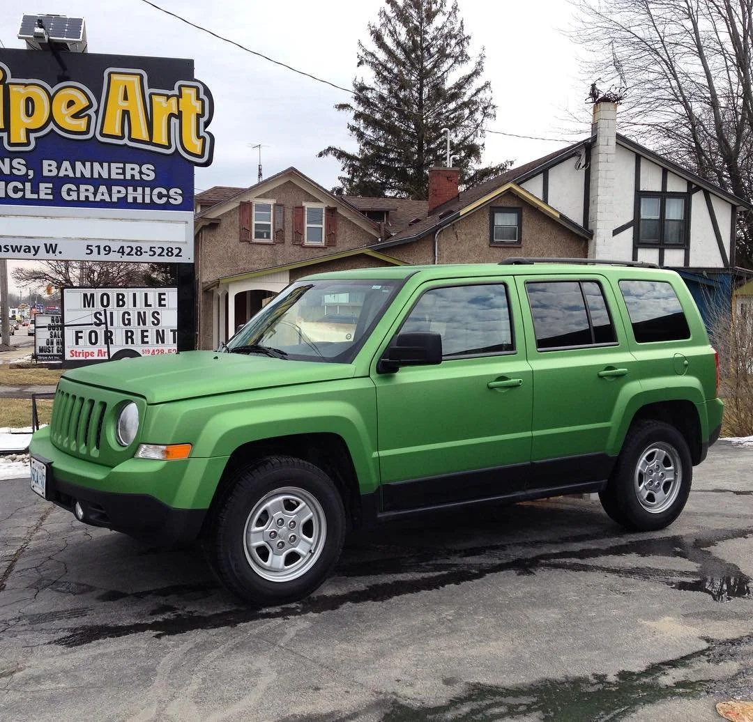 A green Jeep SUV parked on a cracked pavement in front of a row of houses and a sign advertising custom signs and banners.