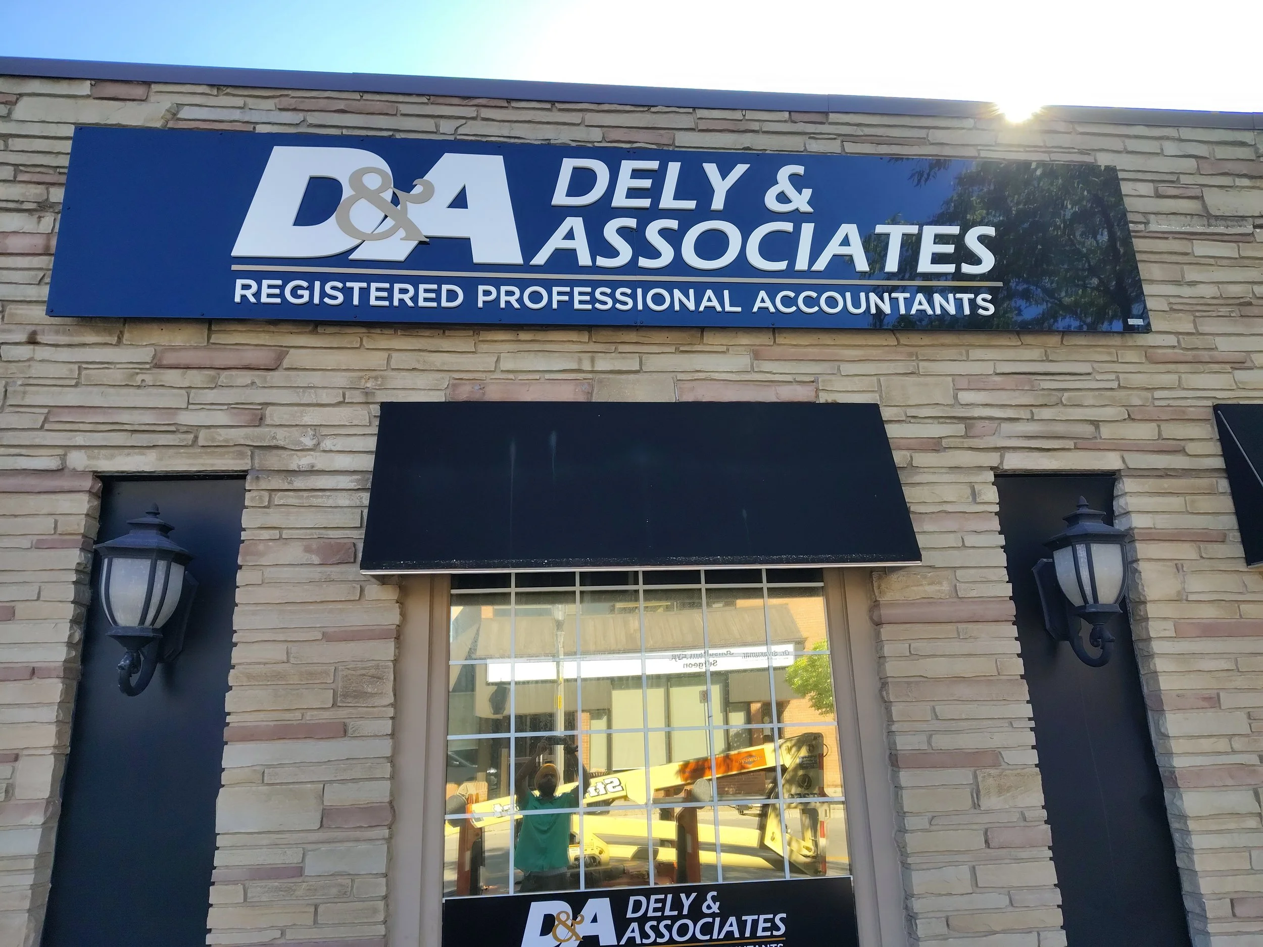 Exterior view of D&A Dely & Associates accounting firm with a blue sign, brick wall, two black outdoor lanterns, and a window reflecting yellow vehicle and a person taking the photo.