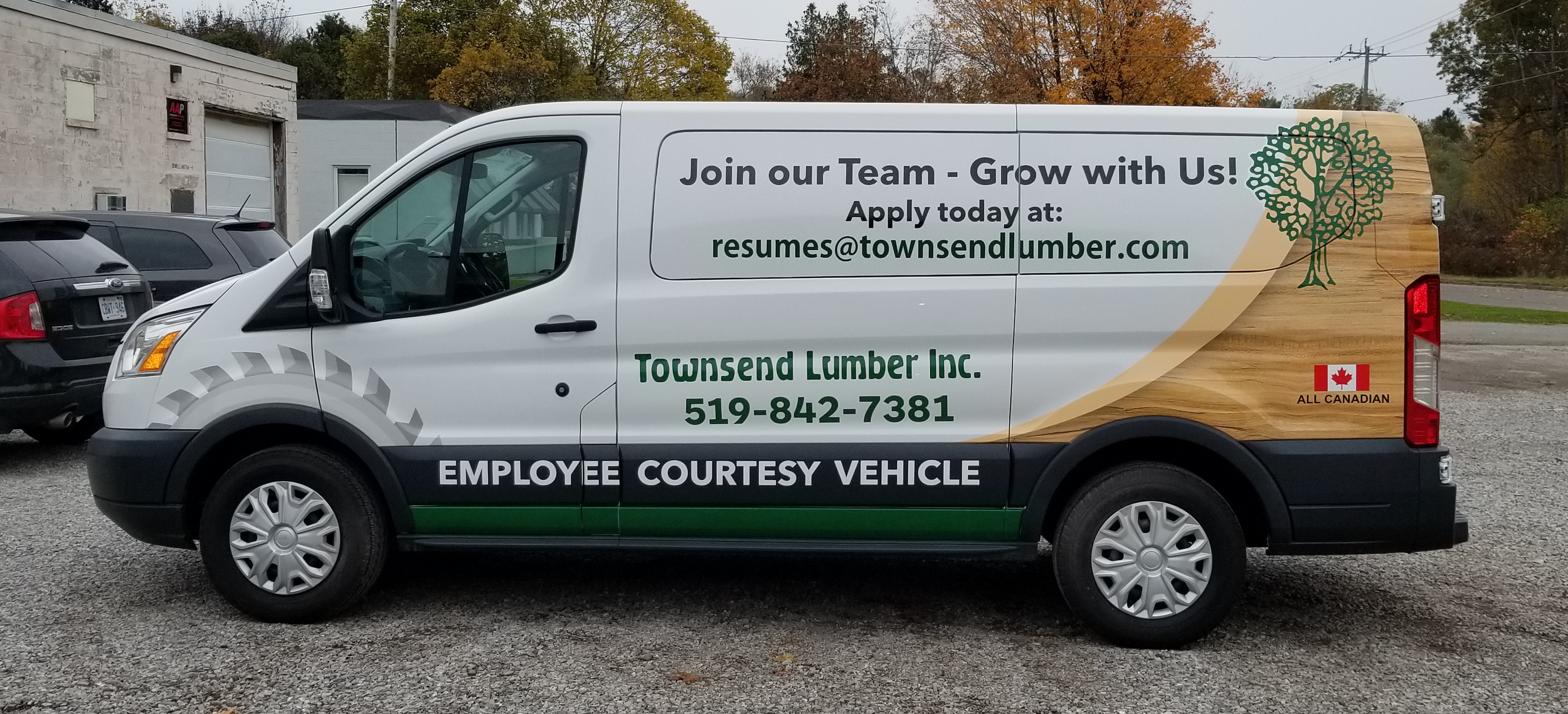 White company van with advertisements for Townsend Lumber Inc., including a green tree logo, a wood grain background, and contact information. The van has a message recruiting employees and an 'All Canadian' sticker with a Canadian flag. It is parked in a lot with other vehicles, and there are trees with autumn foliage in the background.