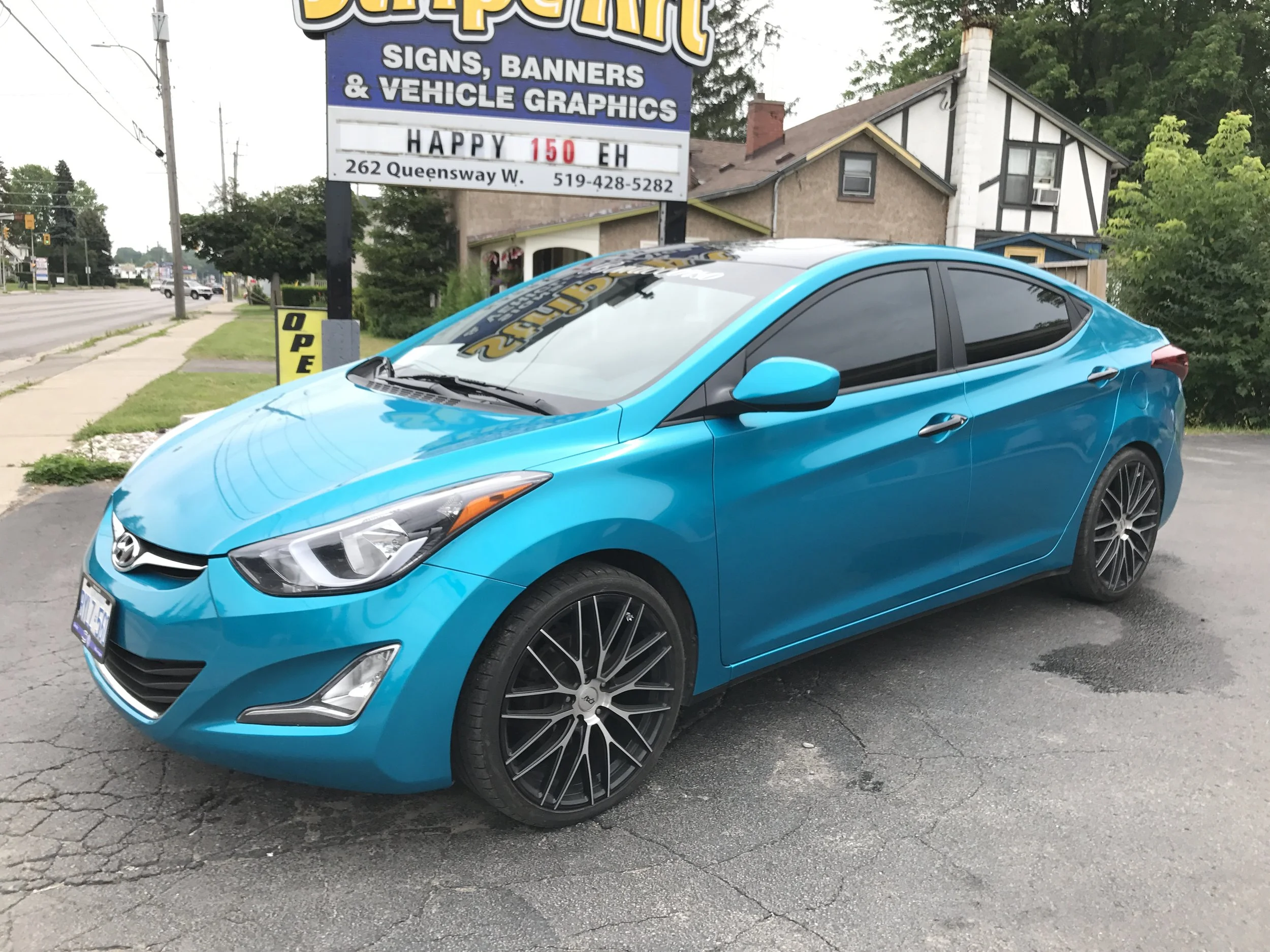 A blue sedan parked in a lot near a sign for a sign and banner shop, with a house and trees in the background.