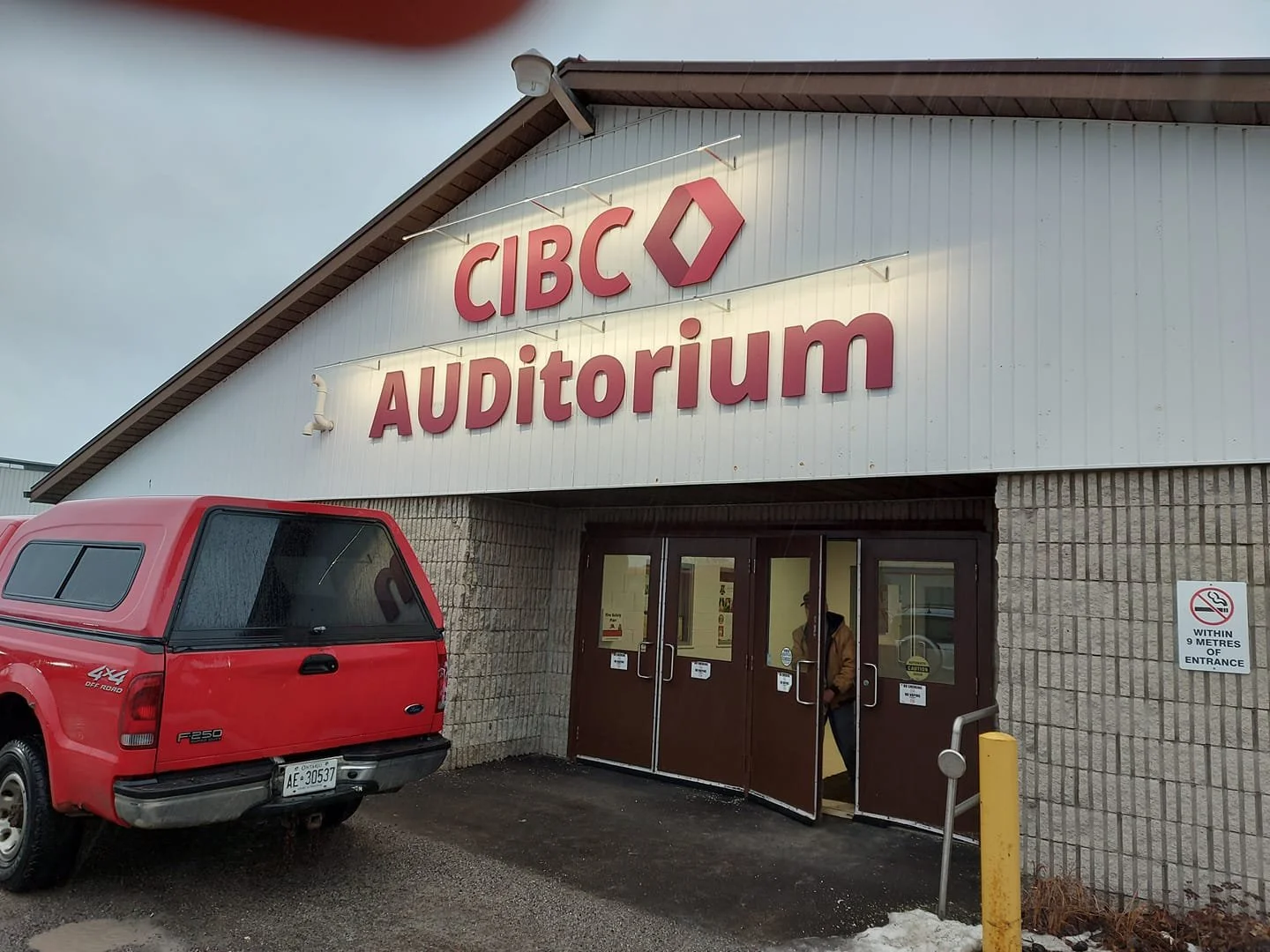 Exterior view of a building with a sign reading 'CIBC Auditorium' and a red pickup truck parked in front. Two people are entering the building through brown doors, and there is a no smoking sign on the wall.