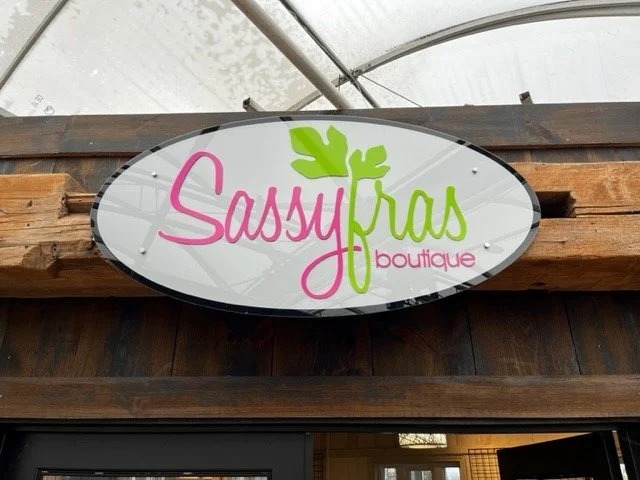 Sign for Sassy Fras Boutique with pink and green lettering and a green leaf logo.