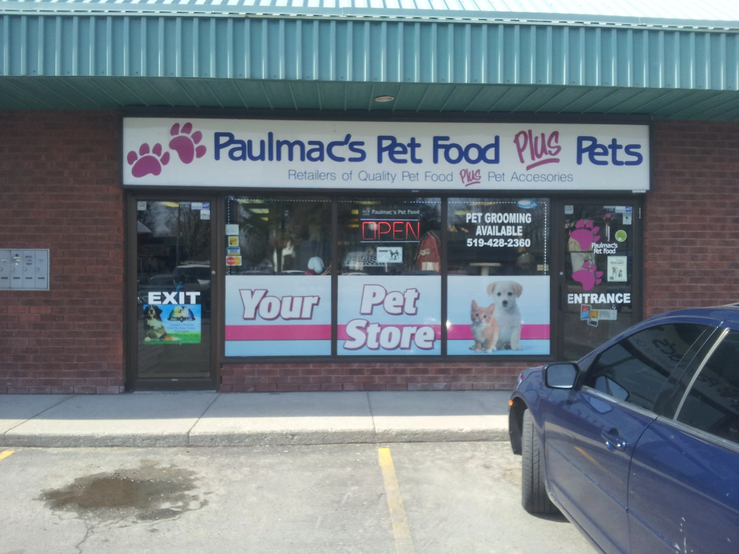 Front view of a pet store called Paulmac's Pet Food Plus Pets, with a large sign displaying pink paw prints and blue text. The store has an 'OPEN' sign in the window, and advertisements for pet grooming and pet food. There is a blue car parked in front of the store and a puddle of water on the parking lot.